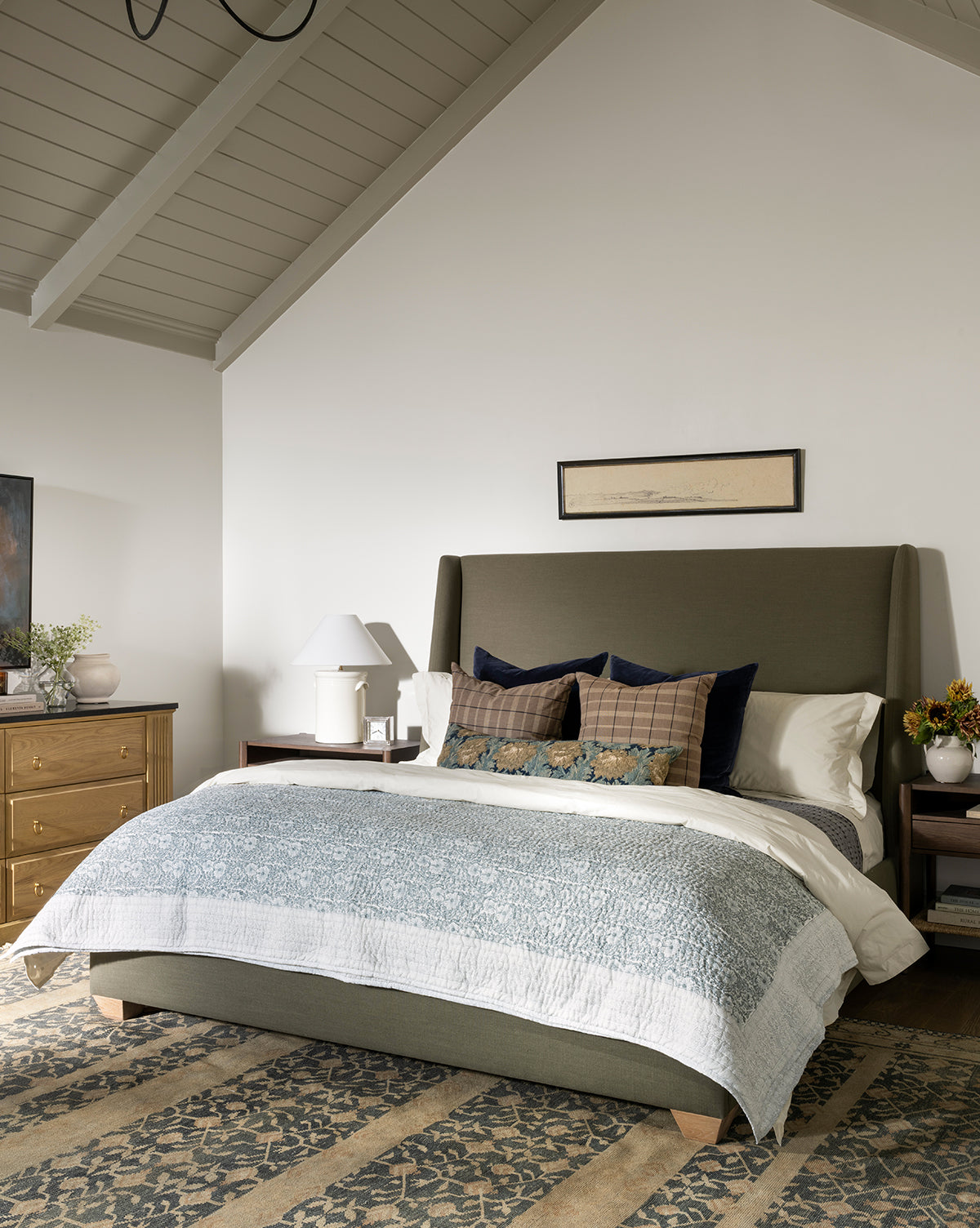 A cozy bedroom features a green upholstered bed, layered pillows, a blue and white quilt with traditional patterns, wooden nightstands with lamps, decorative plants, and the Campanella Hand-Knotted Wool Rug beneath vaulted ceilings with exposed beams, by McGee & Co.