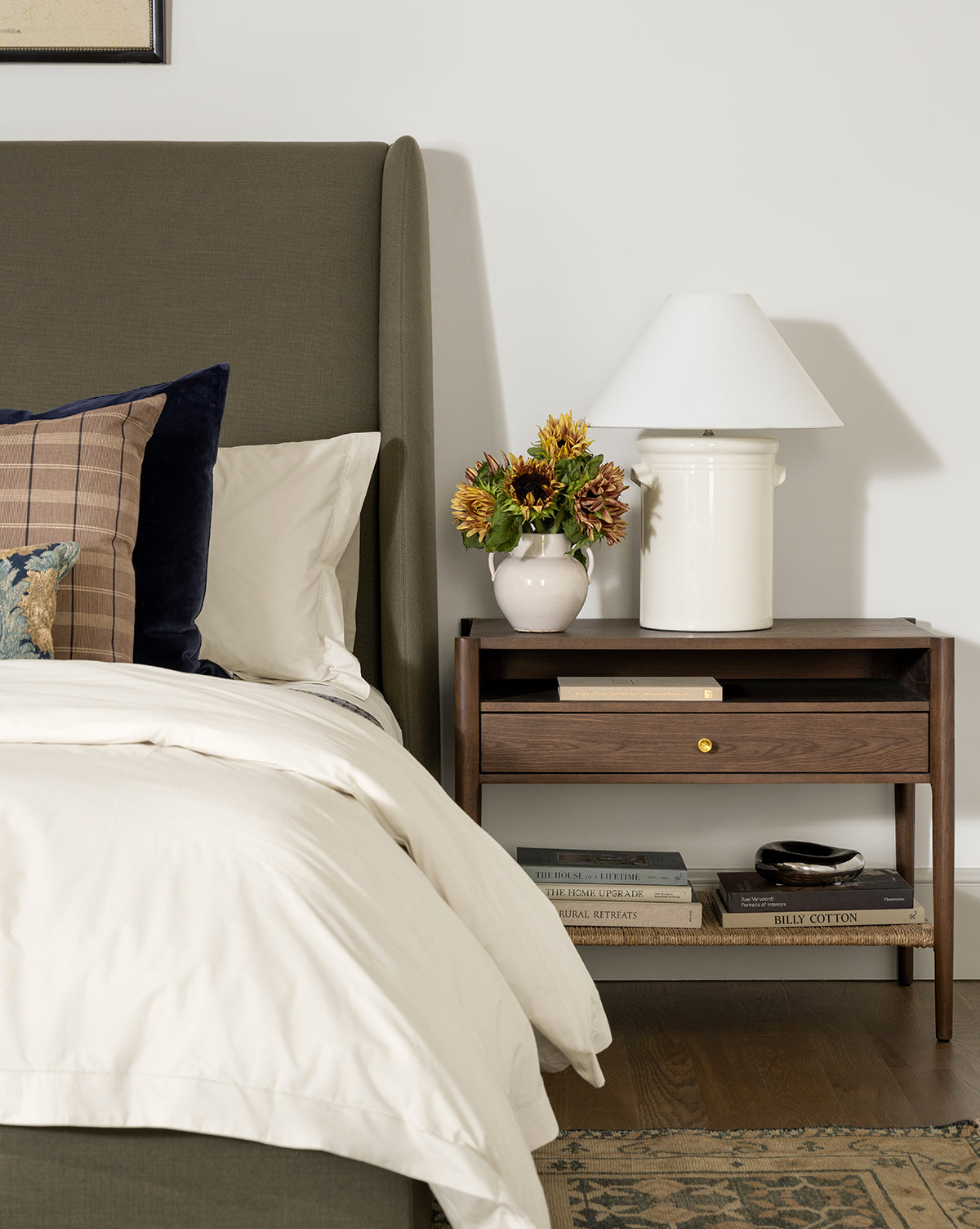A neat bed with white bedding sits beside a wooden nightstand. On the nightstand are the Arles Table Lamp, a vase of flowers, and books. More books are stacked underneath, and part of a rug is visible on the floor, by McGee & Co.