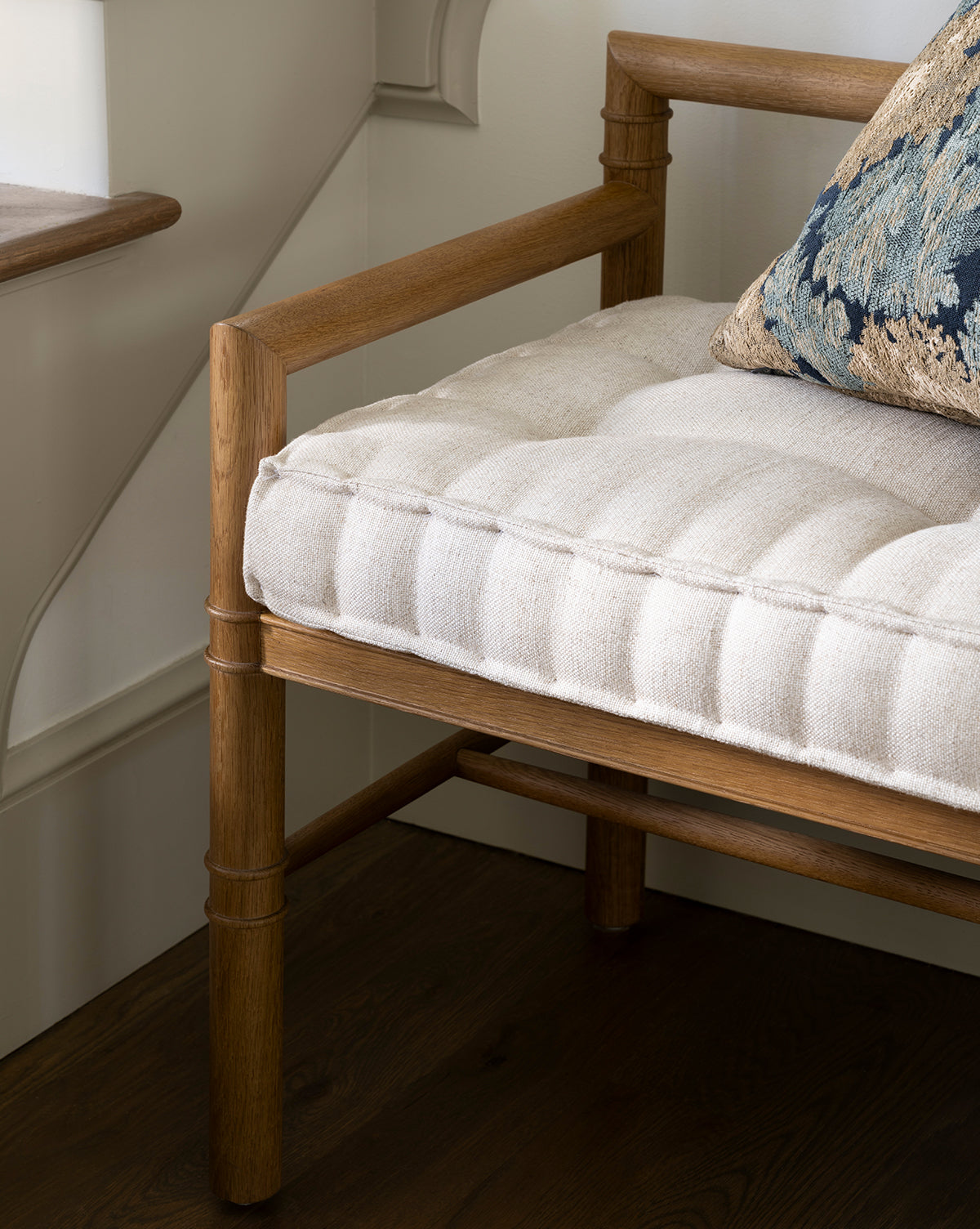A close-up of the Hindley Bench features a light upholstered cushion and a decorative blue patterned pillow, set on a dark wood floor by a window and cream-colored wall, by McGee & Co.