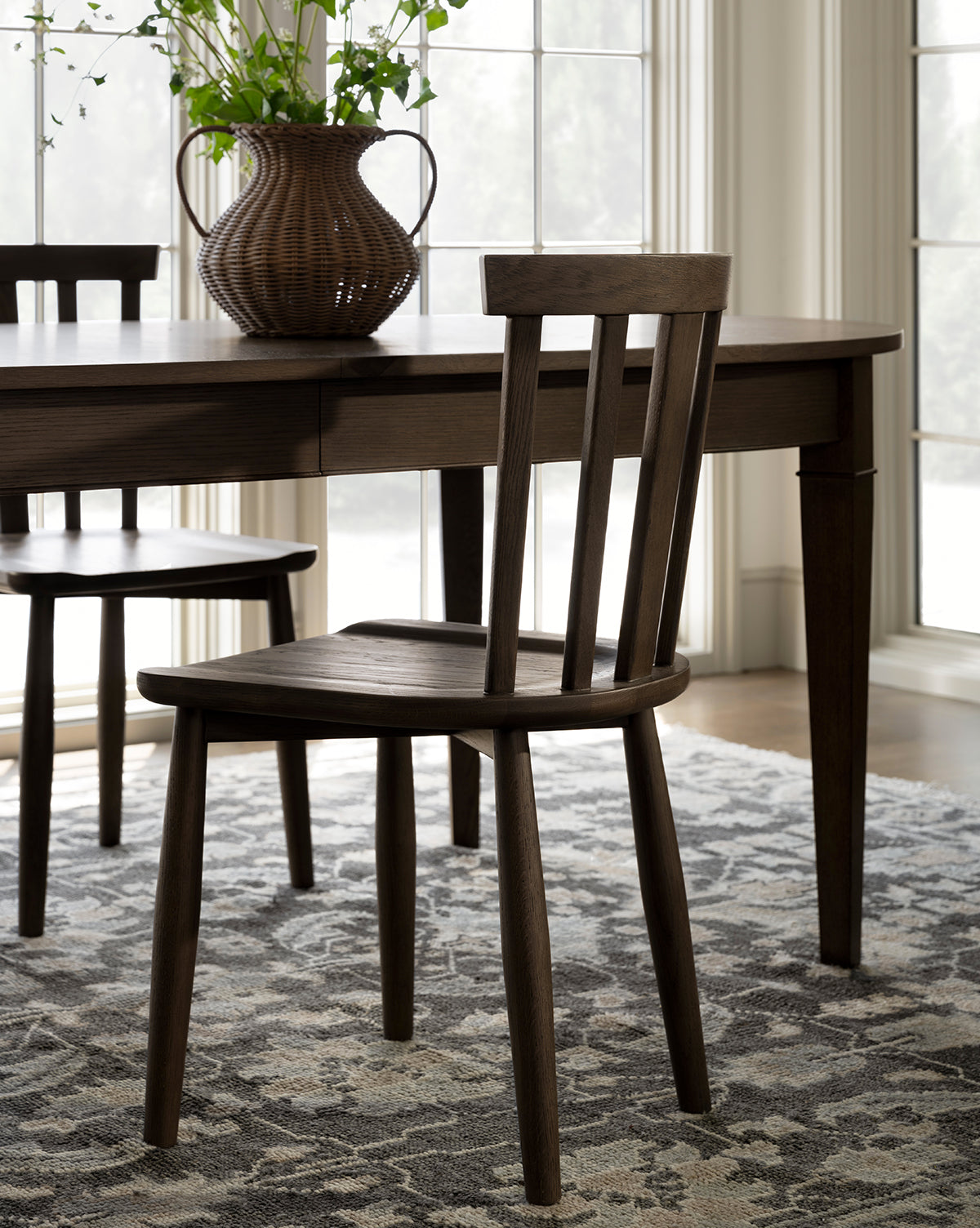 A Hemingway Chair with a slatted back sits on a patterned rug beside a matching table and wicker vase with greenery, illuminated by natural light from large windows, by McGee & Co.