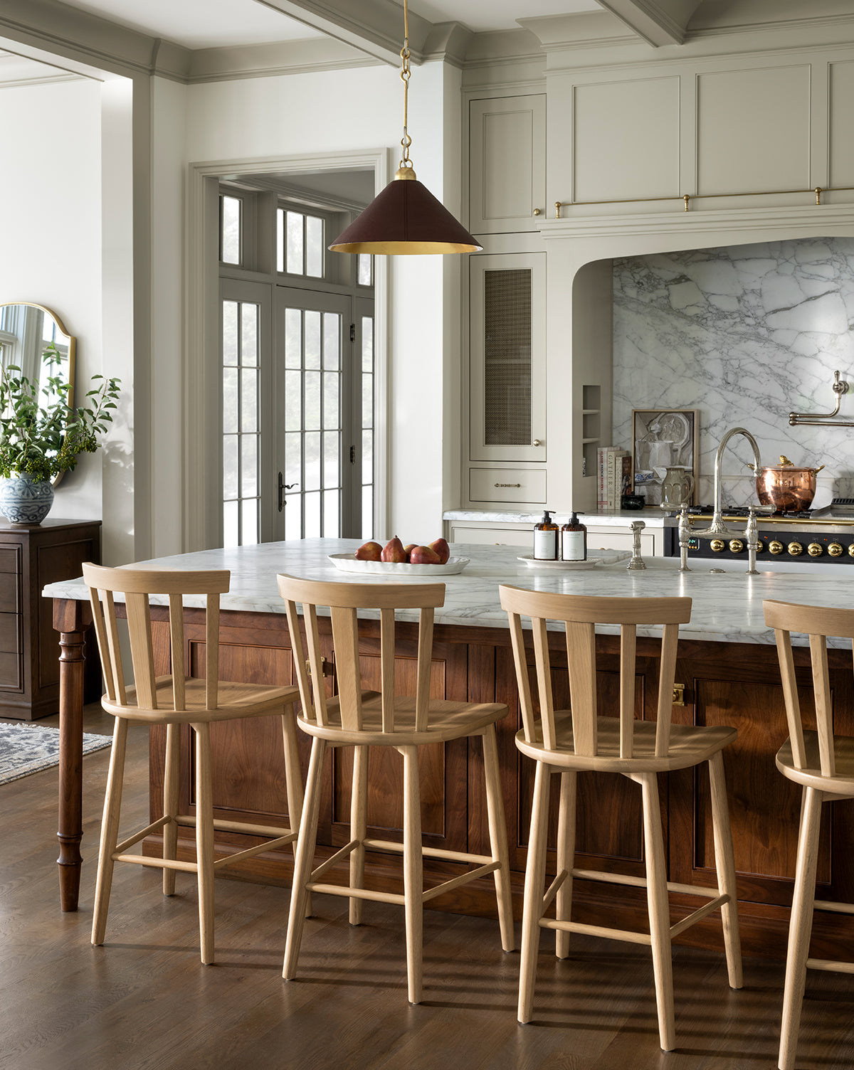 A modern kitchen with a marble island, Hemingway Stools, a copper pendant light, and neutral cabinets. The island features a bowl of apples and soap dispensers, while a potted plant rests near the counters in the background, by McGee & Co.