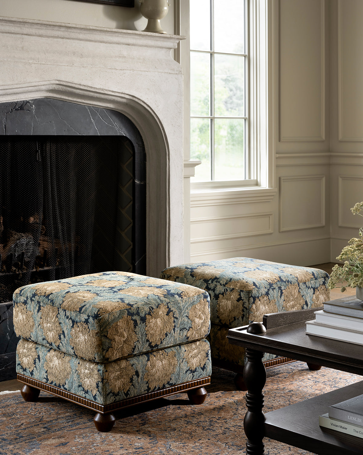 Two Catherine Ottomans, featuring a floral pattern and carved wood base, are placed in front of a fireplace with black marble surround in a bright living room with a window, coffee table, and beige walls, by McGee & Co.