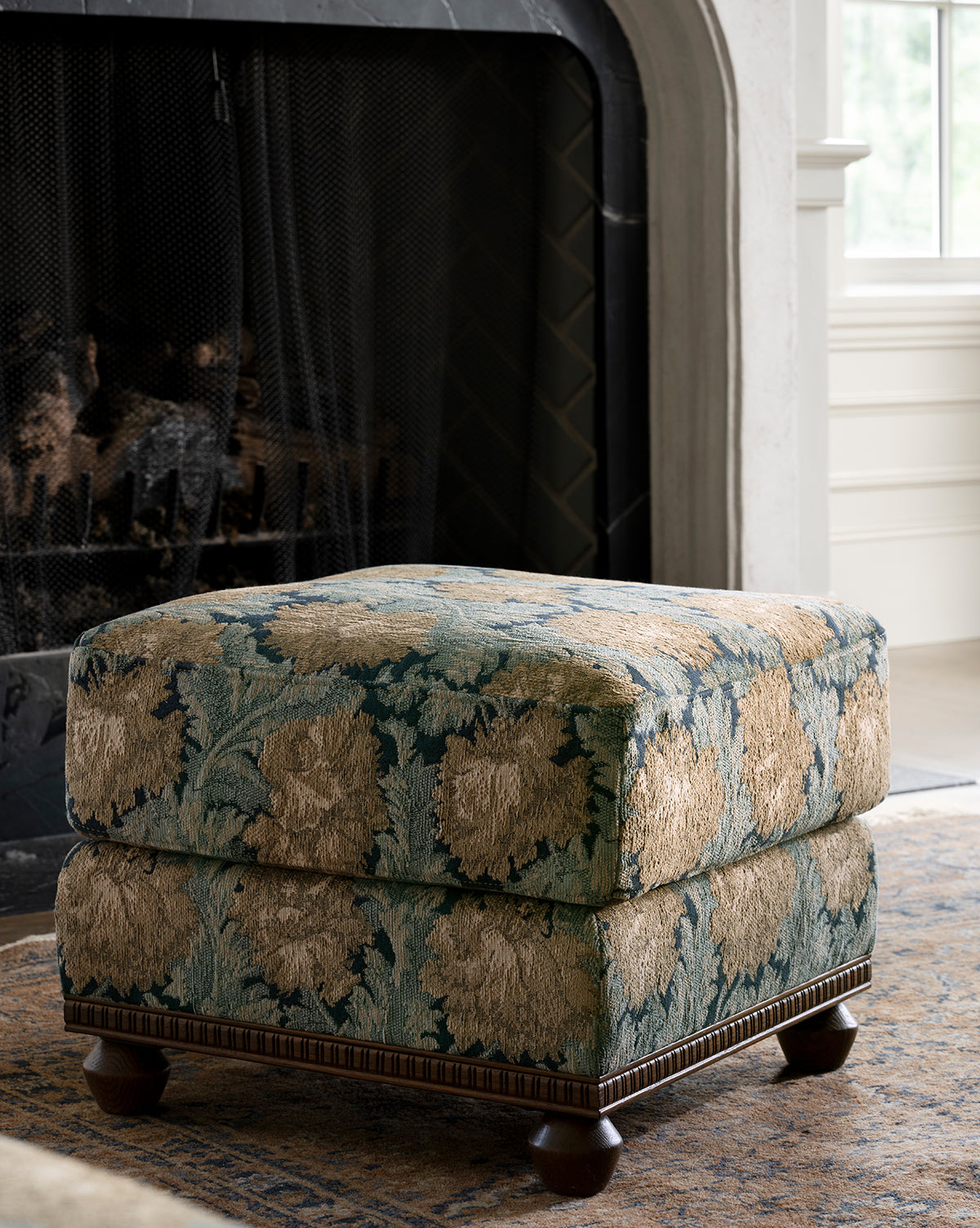 The Catherine Ottoman, featuring a carved wood base and floral upholstery, sits on a rug in front of a fireplace with a black screen in a bright, elegant living room, by McGee & Co.