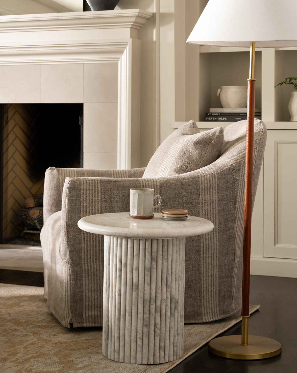 A cozy Trudeaux Slipcover Swivel Chair in beige stripes sits near a round marble-topped side table with a mug and book. A modern floor lamp stands nearby, with a fireplace visible in the background, by McGee & Co.