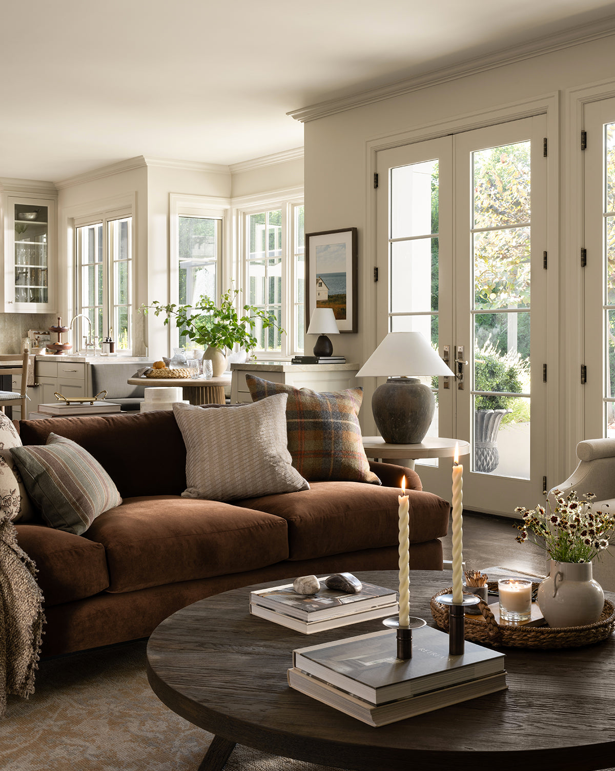 A cozy, sunlit living room features a customizable Lucille English Roll Arm Sofa 90.5 with down wrapped cushions and patterned pillows, a round wooden coffee table with candles and books, large windows, French doors to the garden, and the kitchen in view, by McGee & Co.