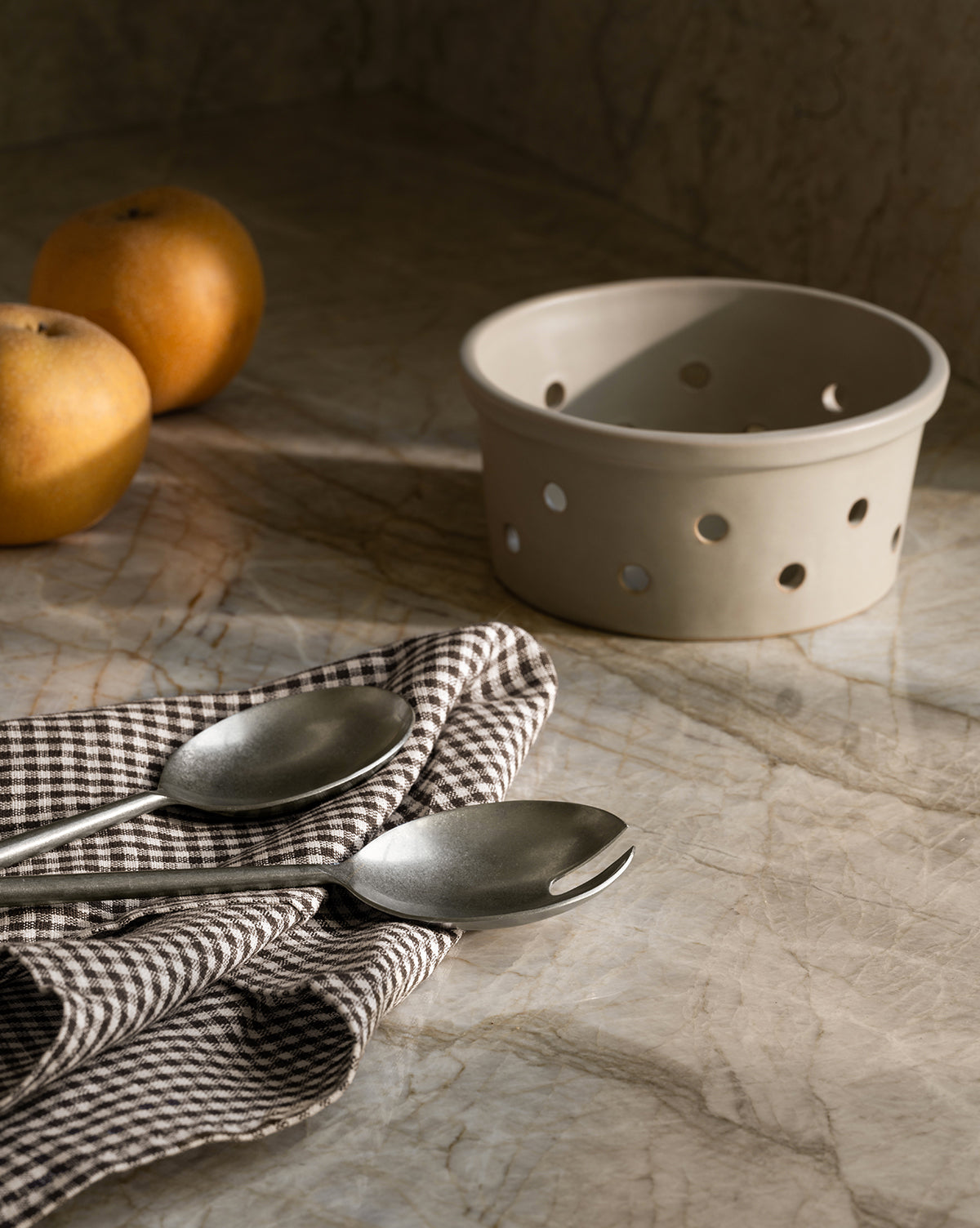 Two metal spoons rest on a brown checkered cloth atop marble. Beside them are two yellow apples and the Coen Berry Bowl, a stoneware bowl with round holes and a matte finish. Soft light casts gentle shadows over the scene, by McGee & Co.