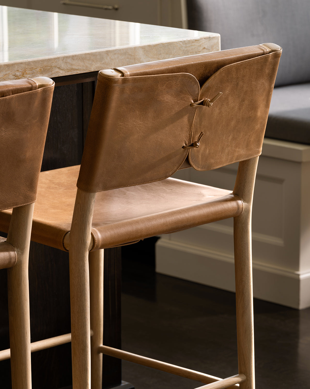 The Danson Counter Stool features a light wooden frame, tan leather seat and backrest with leather lacing, and minimalist design, shown beside a marble countertop in a stylish kitchen by McGee & Co.