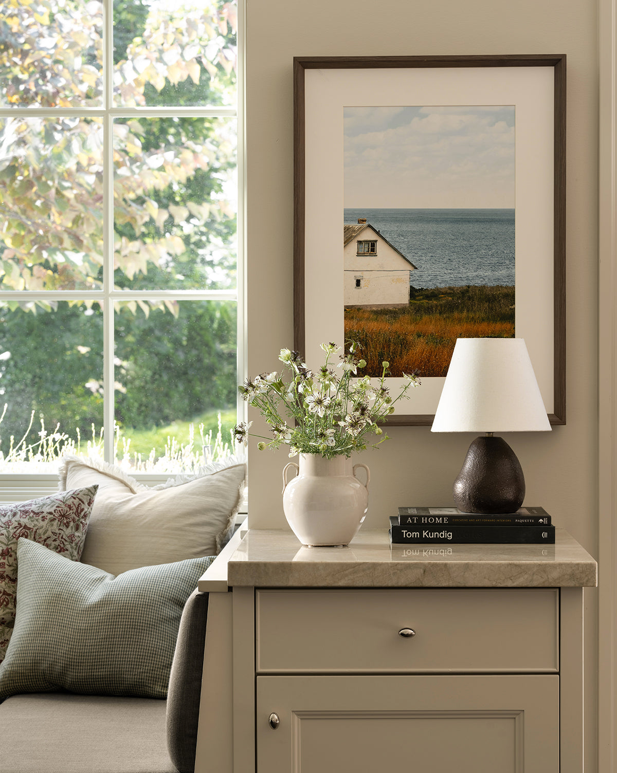 A cozy corner features a cushioned bench by the window, a vase of white flowers, books on a beige cabinet, and the Vedruna Table Lamp with its brown ceramic base and tapered white shade beneath a coastal house photo by McGee & Co.