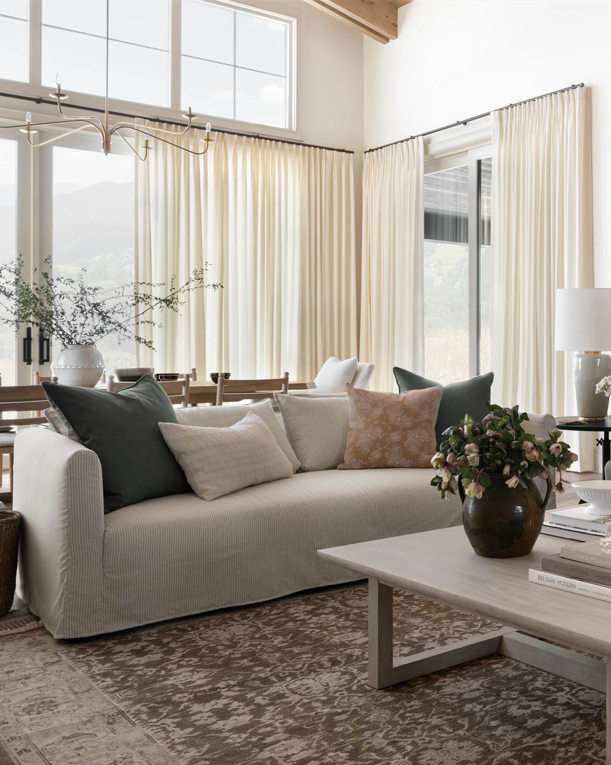 A cozy living room showcases the Rowe Fine Furniture, Inc. Iman Slipcover Sofa (90-96) with throw pillows, a patterned rug, a wooden coffee table topped with books and a vase, large cream-curtained windows, and soft natural light streaming in—McGee & Co.