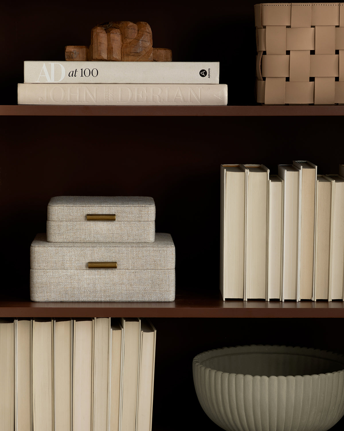 A bookshelf displays neatly arranged books, the Architectural Digest at 100 coffee table book, two stacked fabric boxes with brass handles, a sculpted wooden hand, and a ribbed bowl—timeless decor worthy of Architectural Digest.