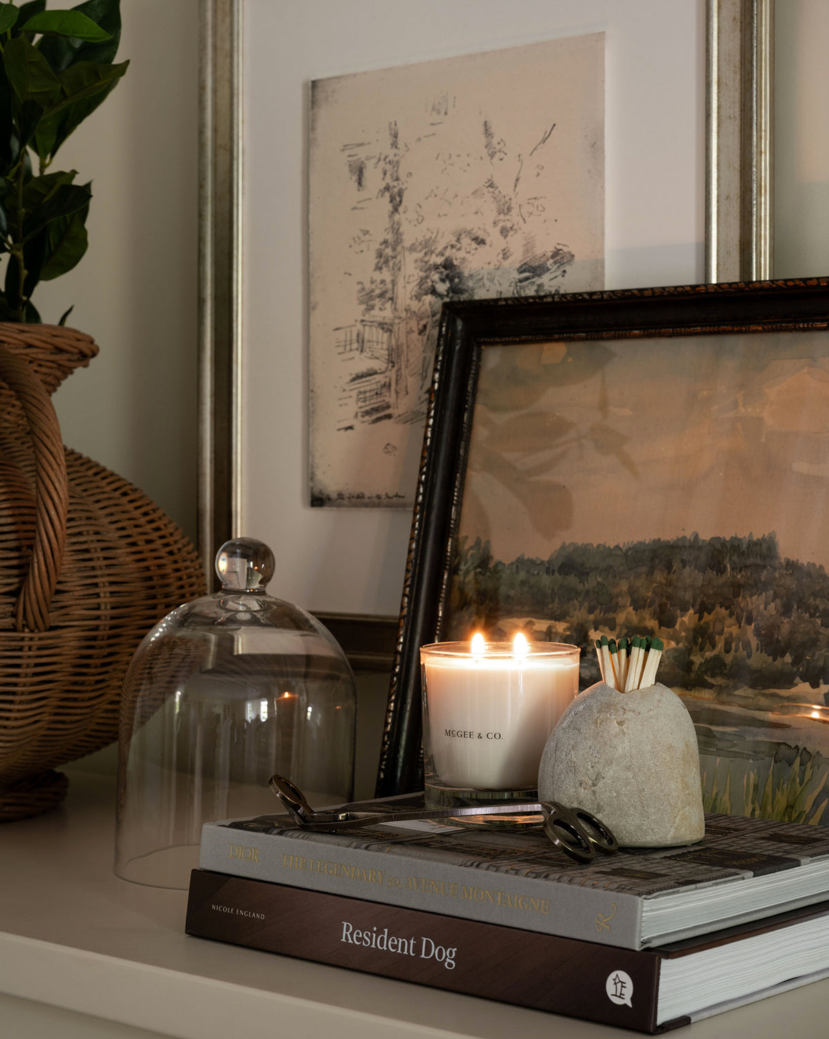A cozy scene with a lit Palo Santo & Oakmoss Candle, stone match holder, art books, framed artwork, a wicker vase of greenery, and a glass cloche on the shelf—warm, inviting, and tastefully arranged by McGee & Co.