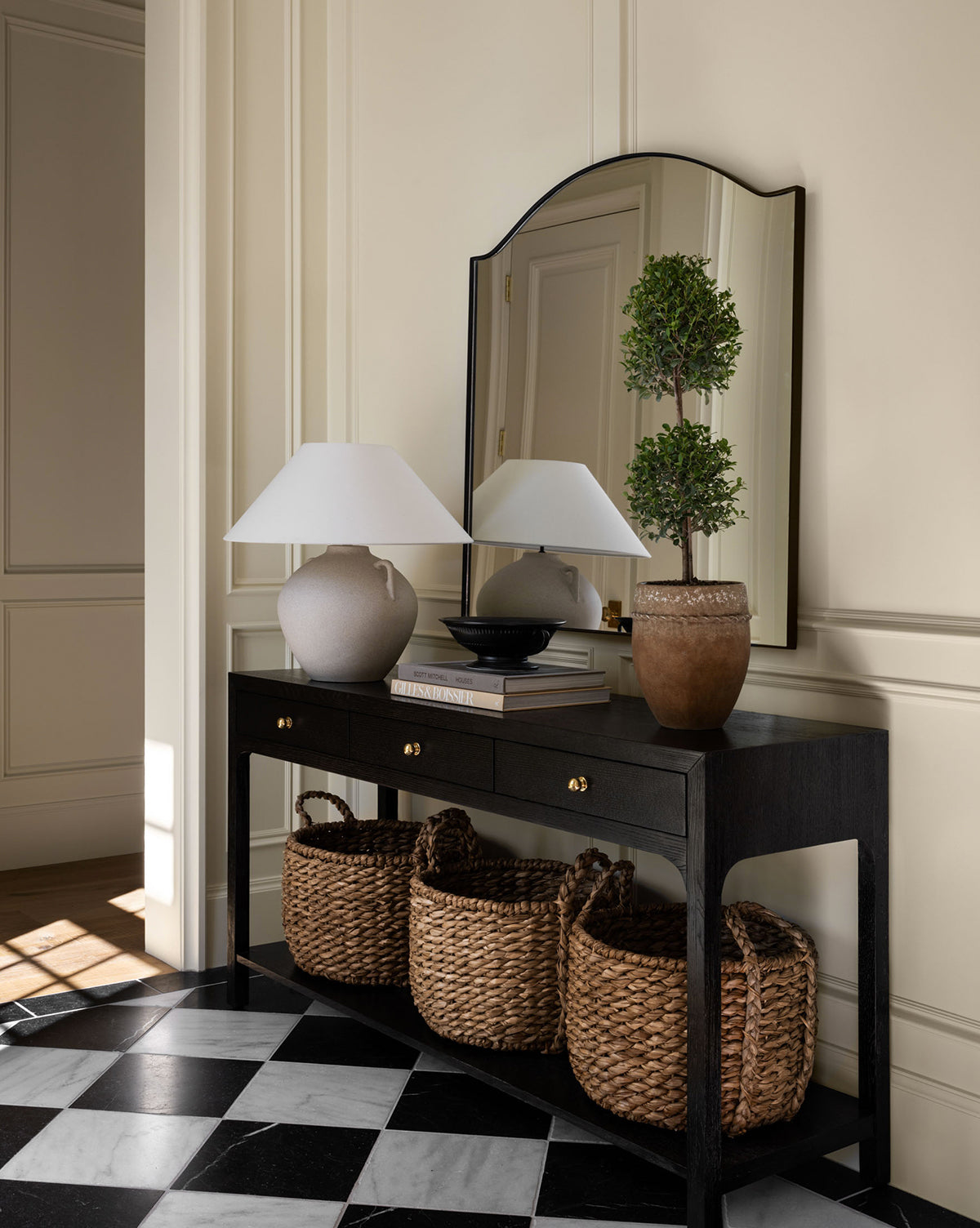 A black console table with drawers displays a white lamp, a bowl, and a potted topiary, with three woven baskets below. The Willson Wall Mirror’s iron frame reflects against cream-paneled walls and checkered black-and-white flooring by McGee & Co.