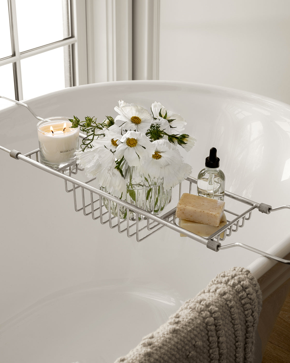 A bathtub with a metal tray holds a lit Salt & Sea Candle, a glass jar of white flowers, a bar of soap, a dropper bottle, and a textured gray towel draped over the edge near a sunlit window, by McGee & Co.