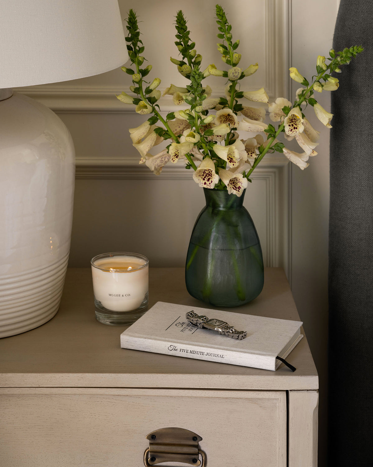 A small beige nightstand holds a green vase of pale yellow flowers, a lit Citrus & Birch Candle, and a closed journal with a silver pen. Nearby are part of a white lamp and a dark curtain, by McGee & Co.