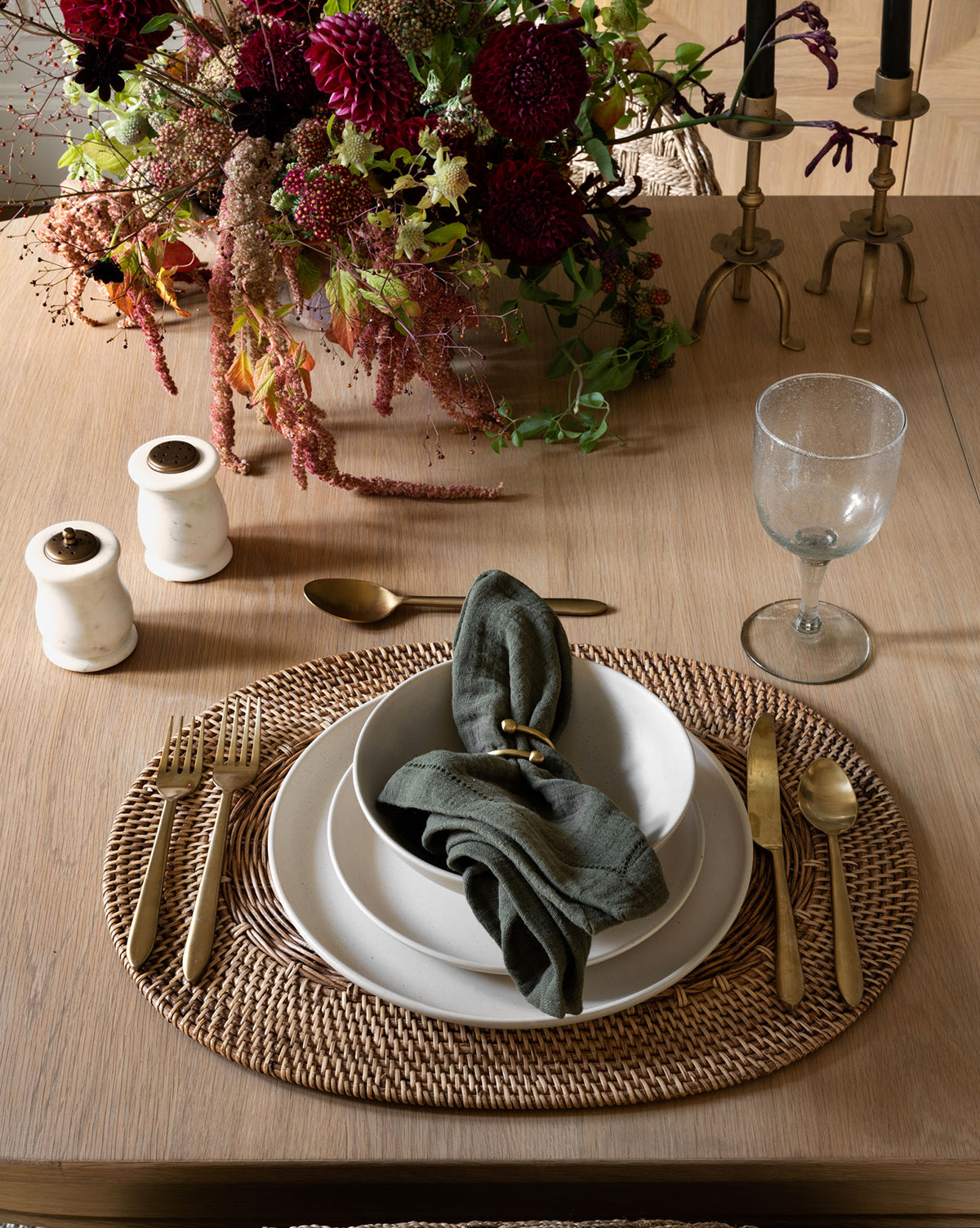 A table setting with gold flatware, white plates, a green napkin, a clear goblet on the Davin Placemat. Salt and pepper shakers, brass candlesticks, and a flower arrangement complete the elegant look, by McGee & Co.