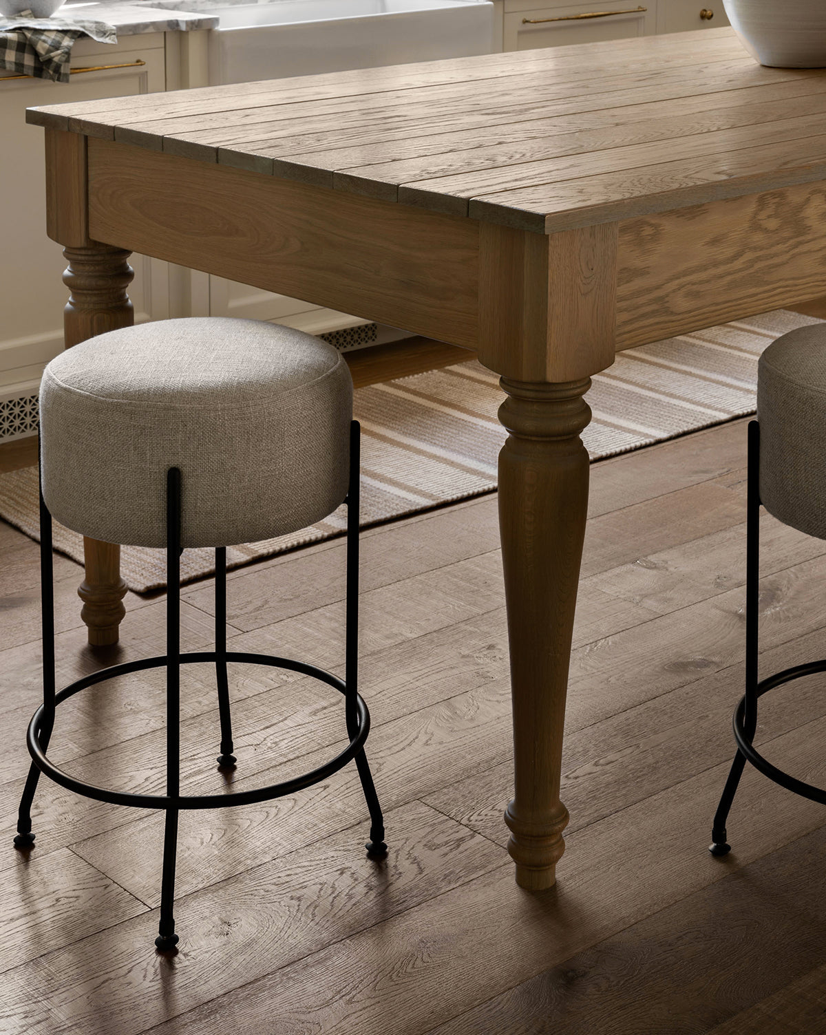 A close-up of a wooden kitchen table with turned legs, two Makerspalm Evander Counter Stools with metal legs, and a striped rug on hardwood in a bright room—ideal for modern kitchen seating. McGee & Co.