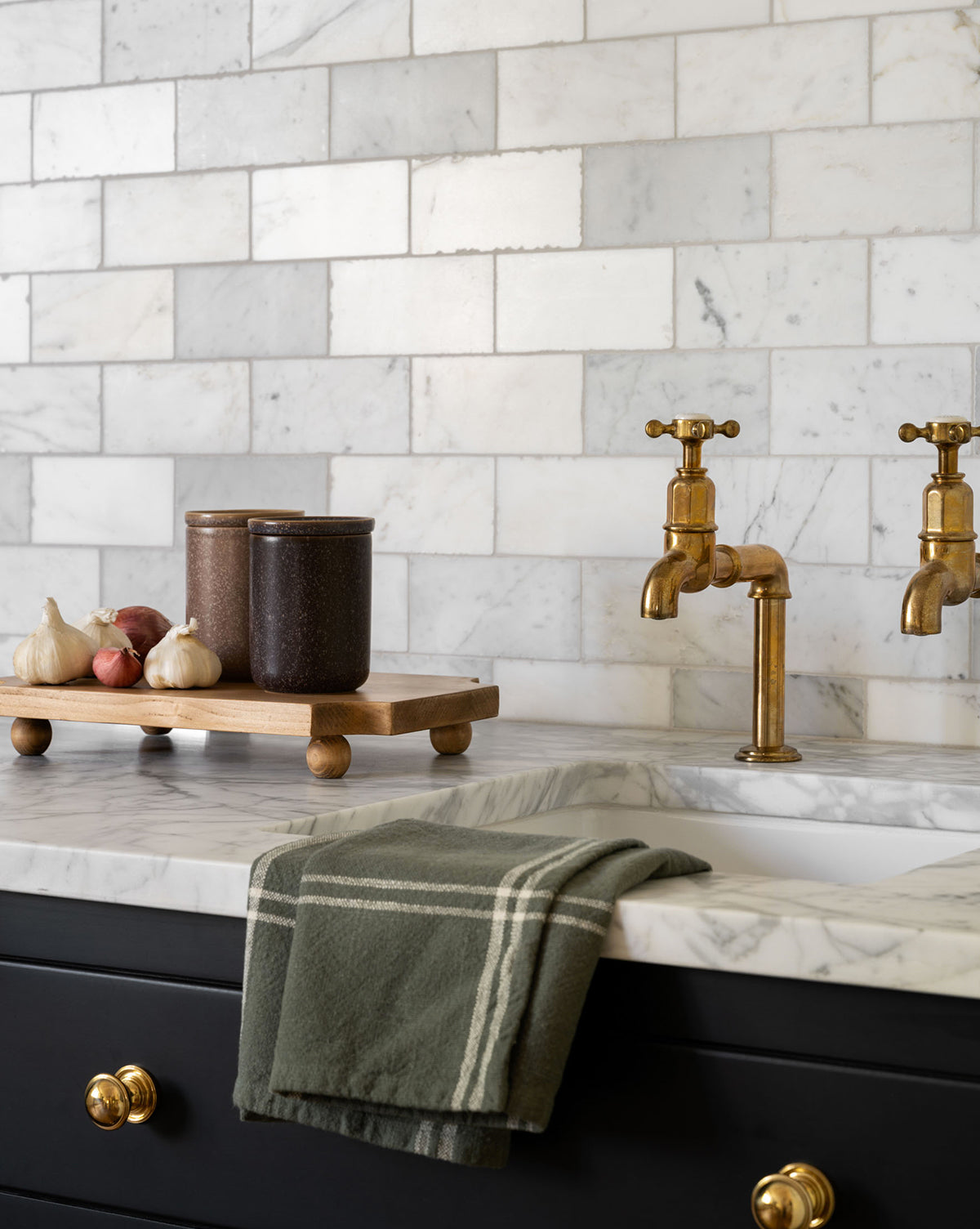 A marble kitchen countertop features a gold faucet, green striped towel, and the Cillian Pedestal Board in pine wood displaying jars and garlic cloves. White marble tile backsplash and gold drawer knobs complete the look by McGee & Co.