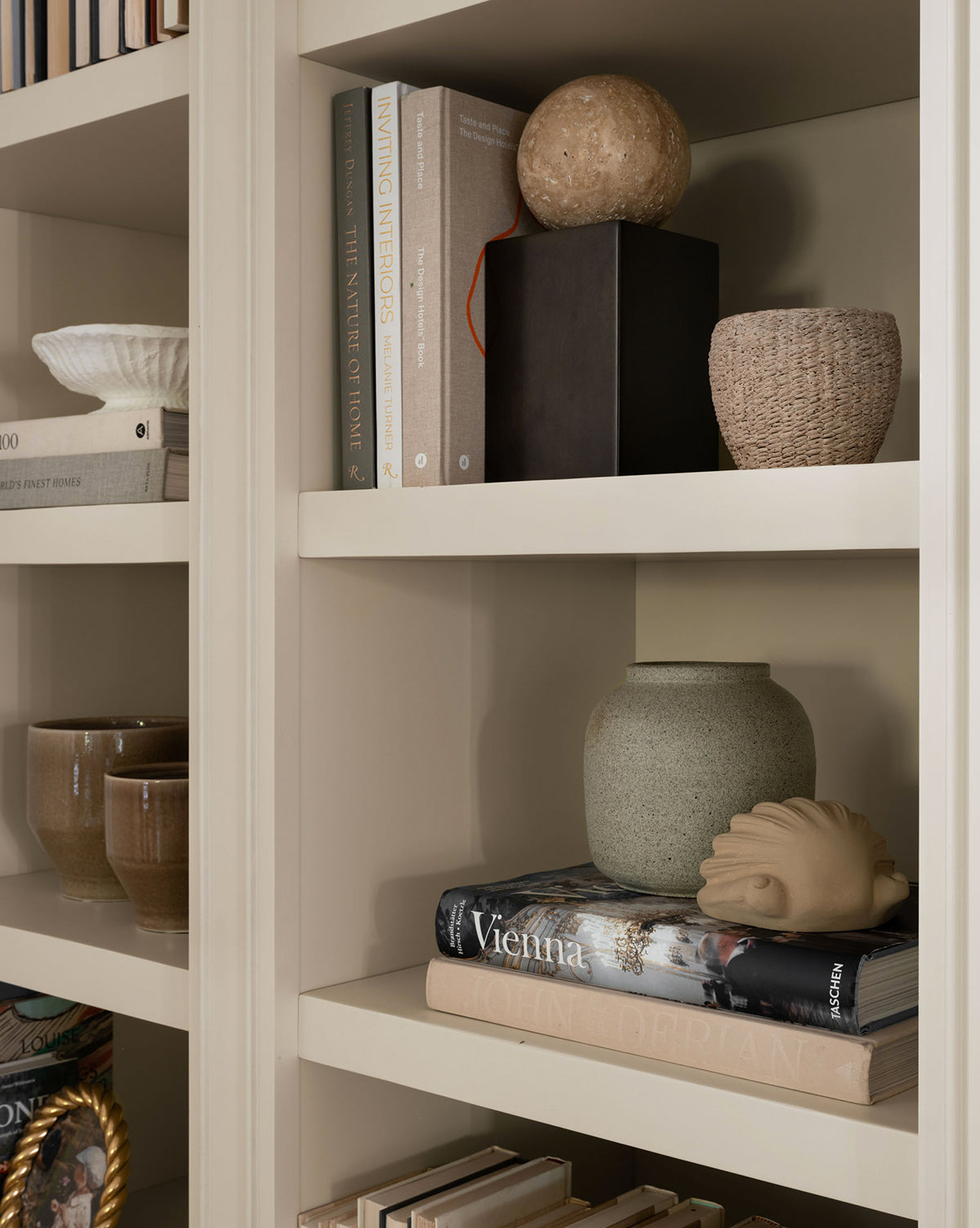 A beige bookshelf displays stacked books, ceramic mugs, a textured vase, the Halston Cement Planter, a cement basket, a round stone, and a decorative scallop shell sculpture—all neatly arranged on its shelves.