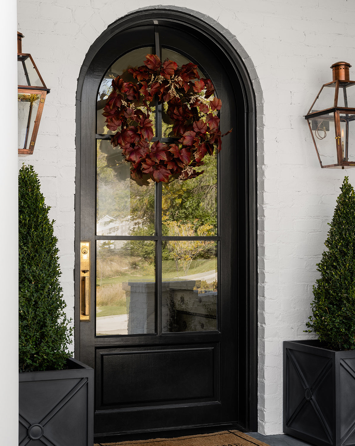 A black arched front door with glass panels displays a Faux Autumn Grapevine Wreath of dark red leaves. Two tall potted topiaries with faux greenery and copper lanterns complete the seasonal decor for the white brick entryway.