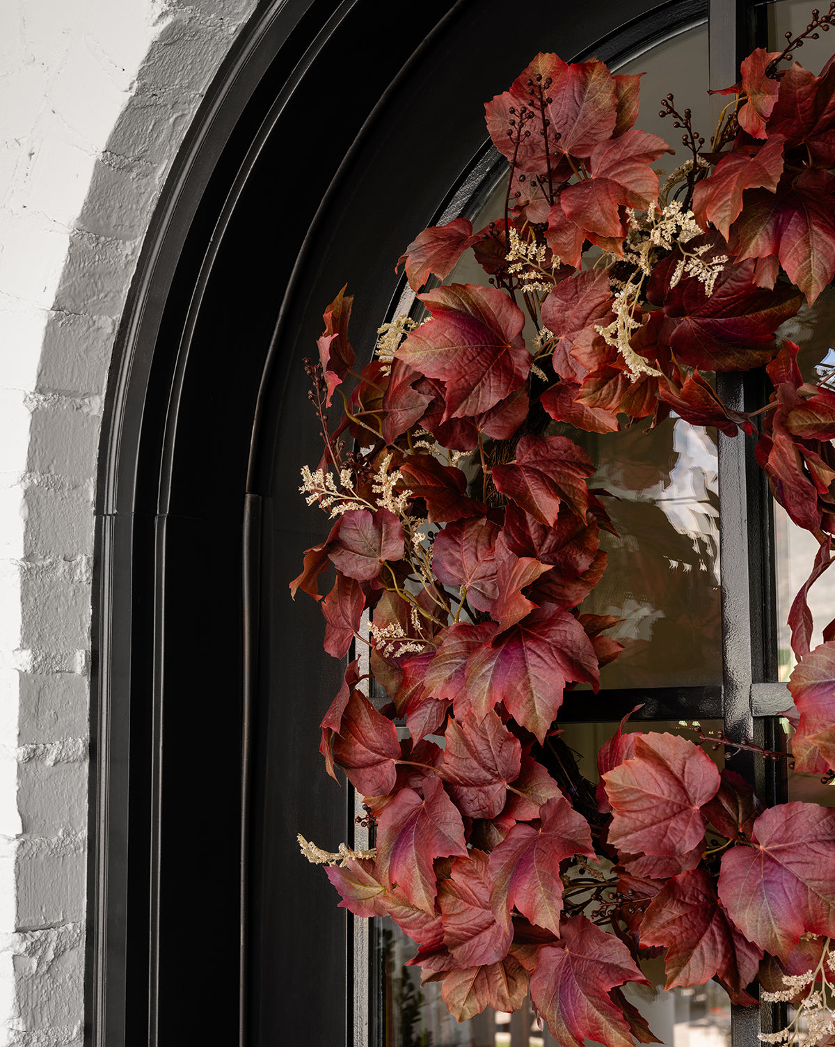 A black arched door features the Faux Autumn Grapevine Wreath, decorated with red and burgundy leaves, faux greenery, and white flowers, offering vibrant seasonal style next to a white brick wall.