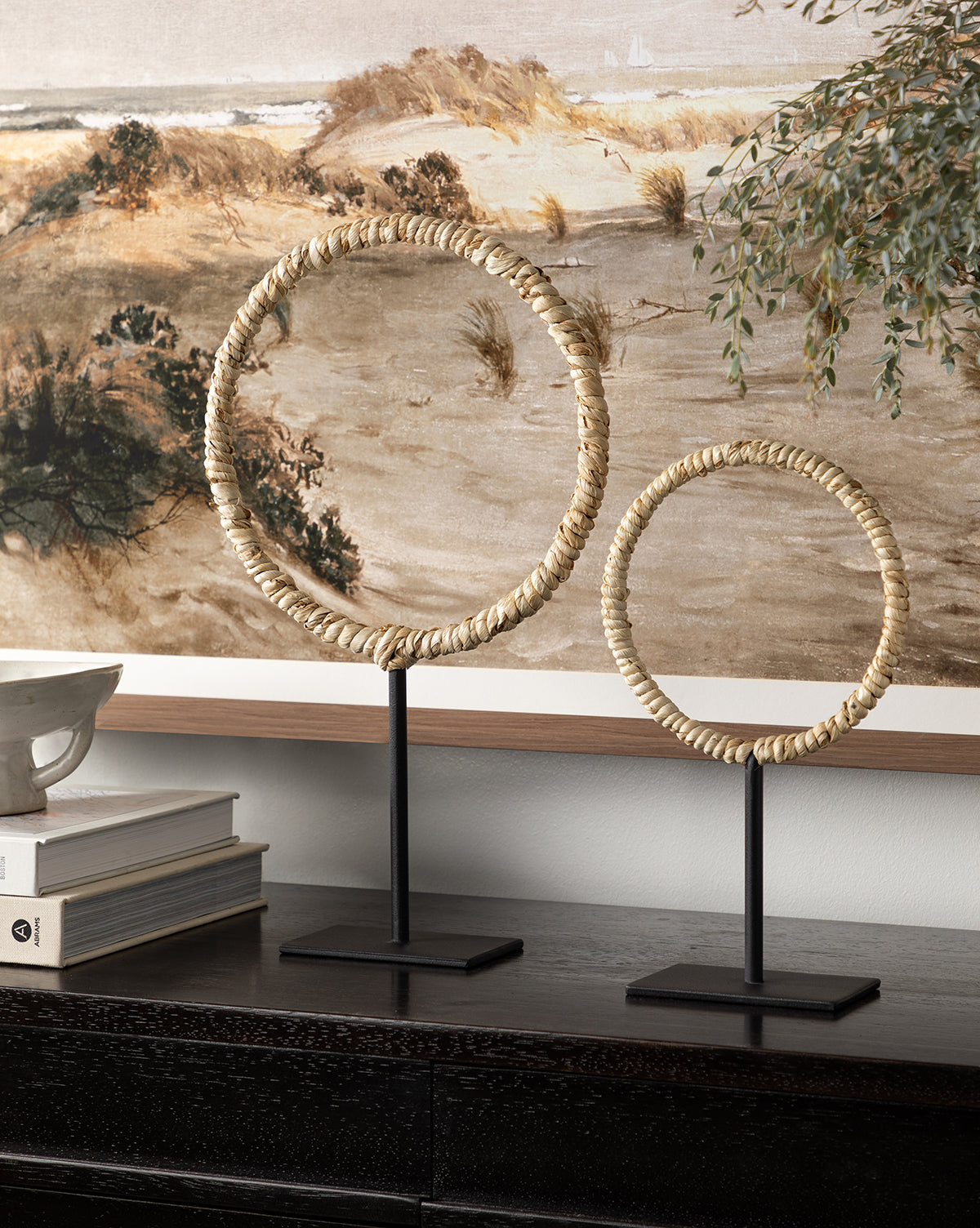 Two Brayden Woven Objects—decorative circular sculptures with a corn husk texture—stand on black metal bases atop a dark wood surface, complemented by a stack of books, ceramic dish, and landscape painting for modern appeal.