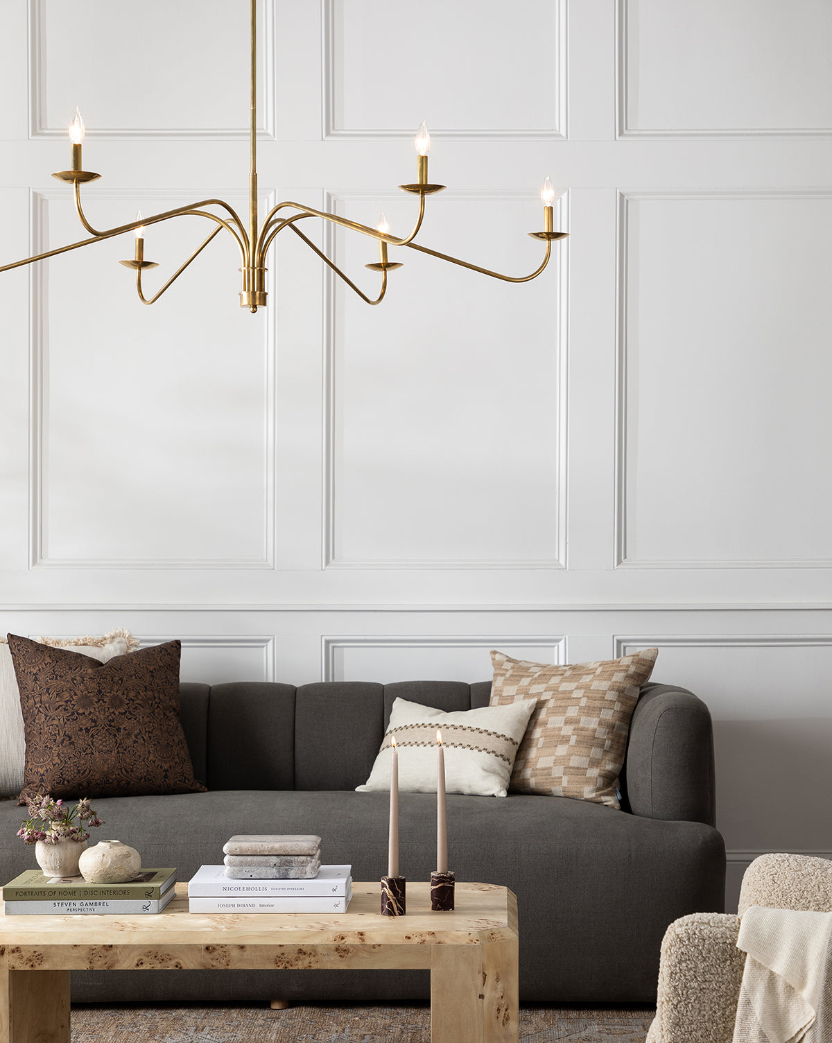 A modern living room featuring the Ines Sofa 90 in gray velvet with patterned pillows, a light wood coffee table with books and candles, and a gold chandelier set against white paneled walls, by McGee & Co.
