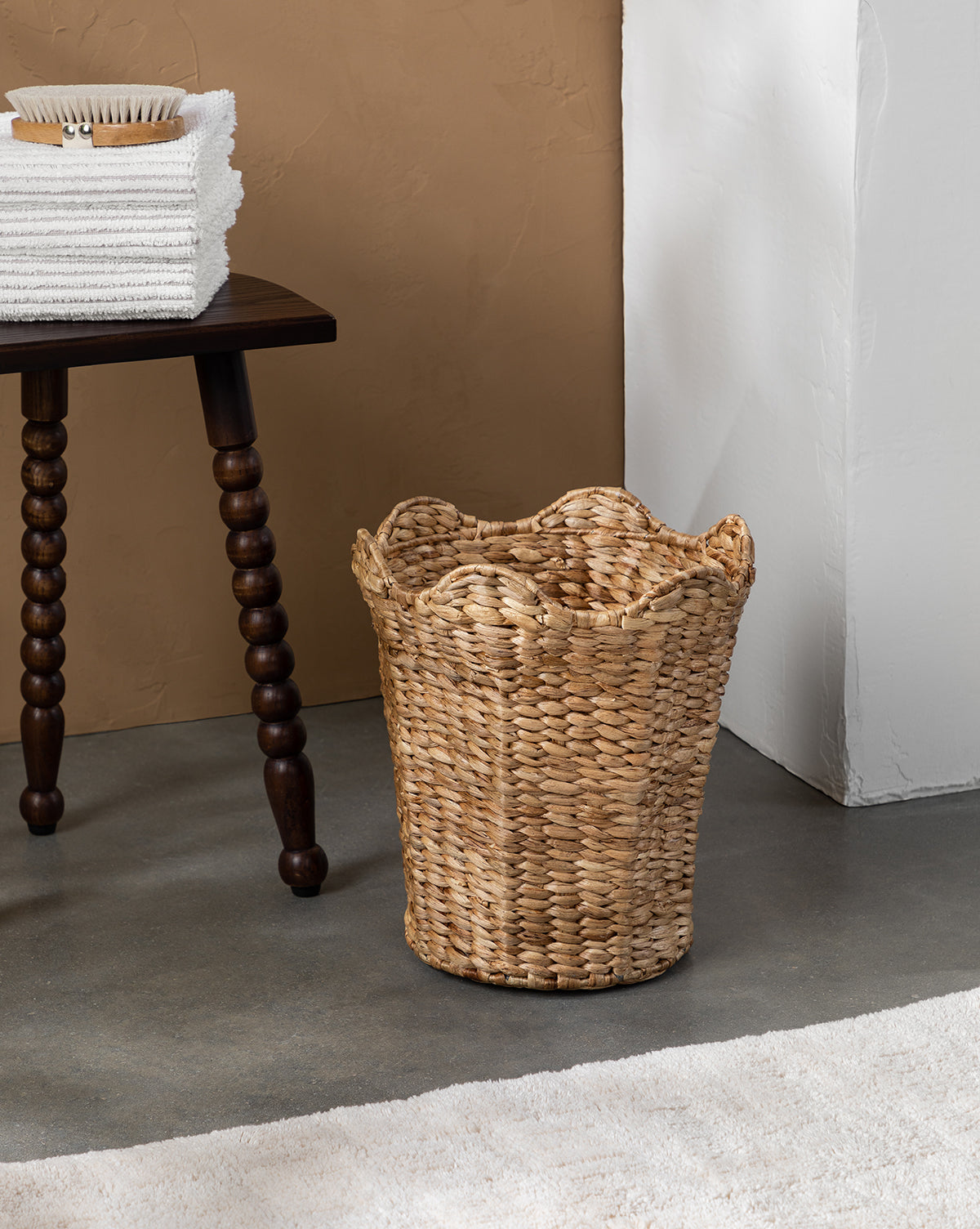 The Tenley Woven Waste Bin with a scalloped rim sits on a concrete floor beside a dark wooden table with folded white towels and a brush. A white rug and textured tan and white walls complete the background.