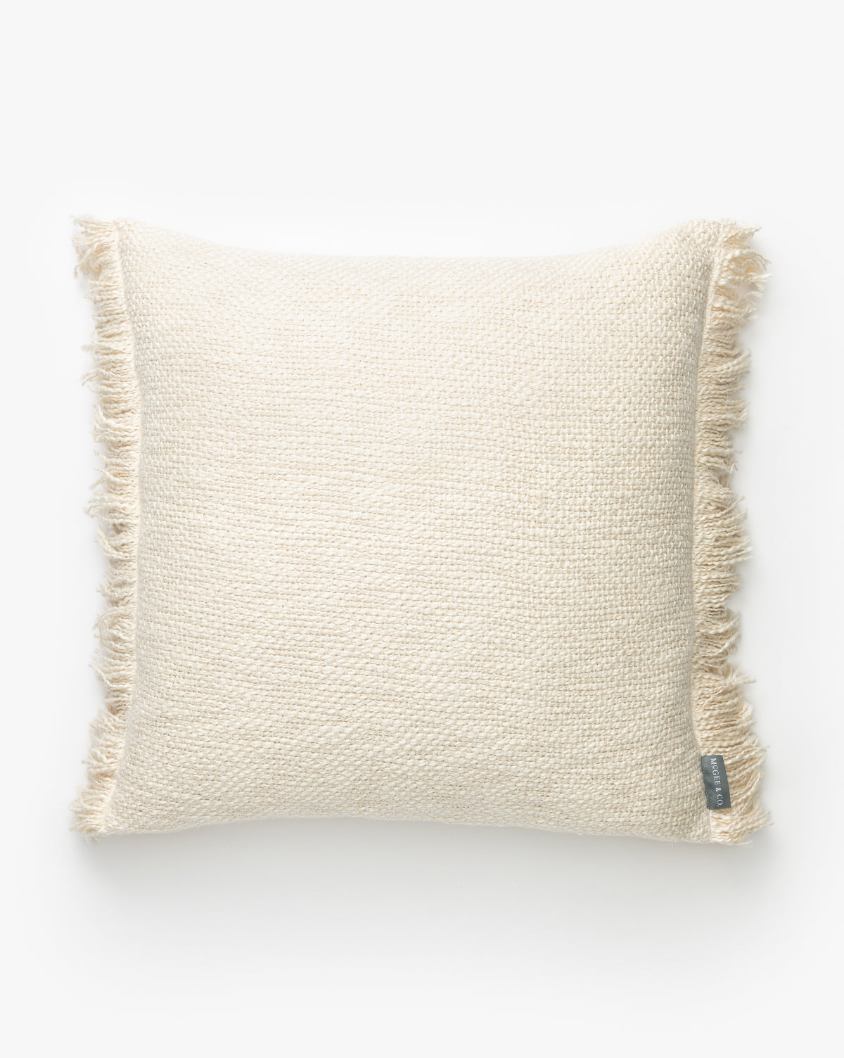 The Aisling Pillow Cover is a square, cream-colored hand-woven pillow cover with a textured design and fringed edges, shown on a plain white background. A small fabric tag appears at the bottom right corner, by McGee & Co.