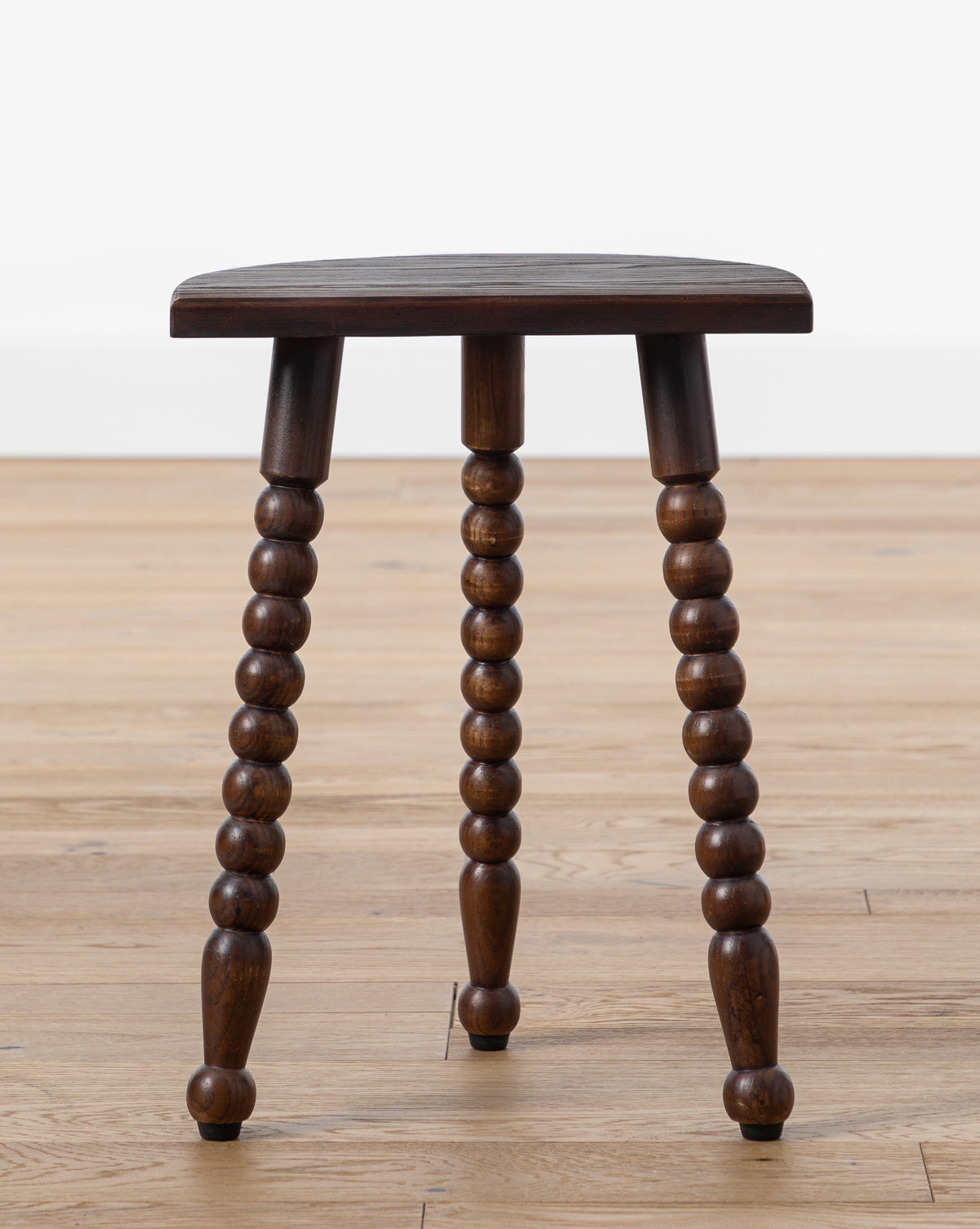 The Alice Wood Stool by Makerspalm is a vintage-inspired ash wood stool with a dark polished seat and turned, beaded legs, shown on a light wood floor against a plain white background by McGee & Co.