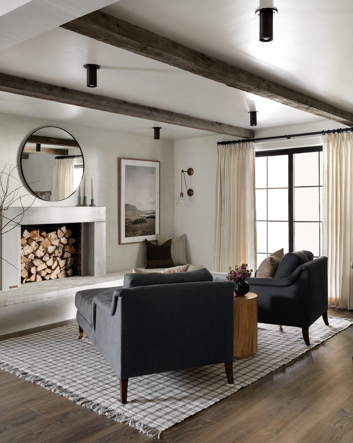 Modern living room with exposed wooden ceiling beams, dark gray sofas, and a Lucille English Roll Arm Chaise Lounge with a down-wrapped cushion, by McGee & Co. A round mirror tops the white fireplace filled with firewood; large window and patterned rug complete the space.