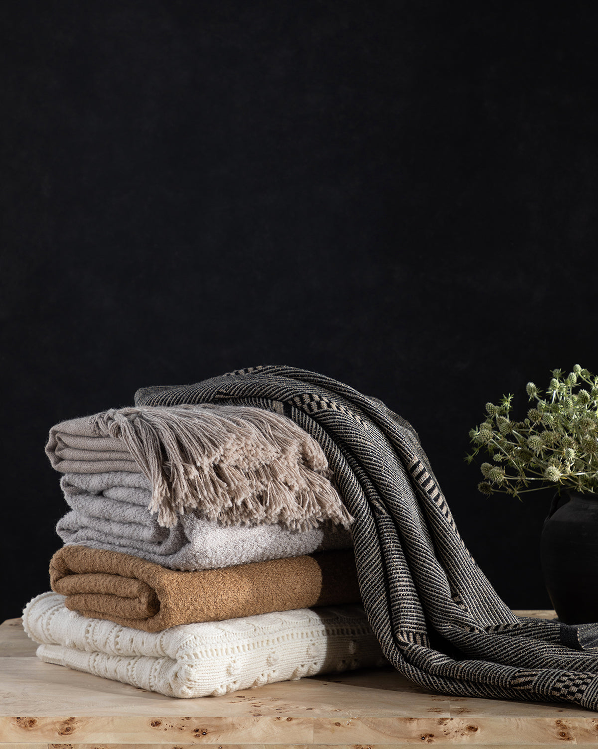 A stack of cozy, neutral-toned blankets sits on a wooden surface, with the Anwar Woven Throw—featuring black-and-white stripes—draped over the side. A small potted plant in a dark vase adds charm against the black background.
