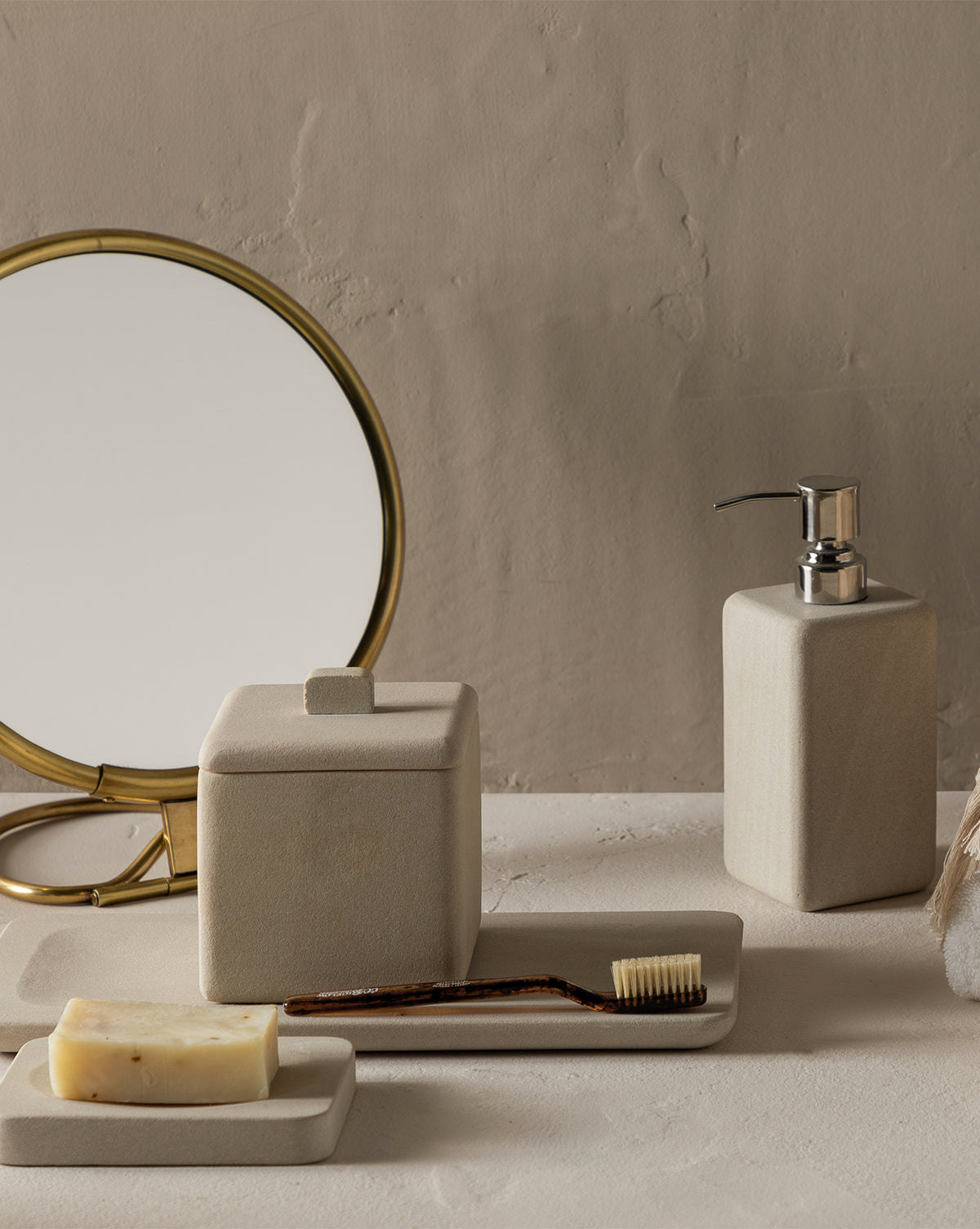 A minimalist bathroom setup with a gold-framed round mirror, the Amada Sandstone Bath Collection, a bamboo toothbrush, and a beige soap dispenser adds an organic touch to the countertop against a textured beige wall.