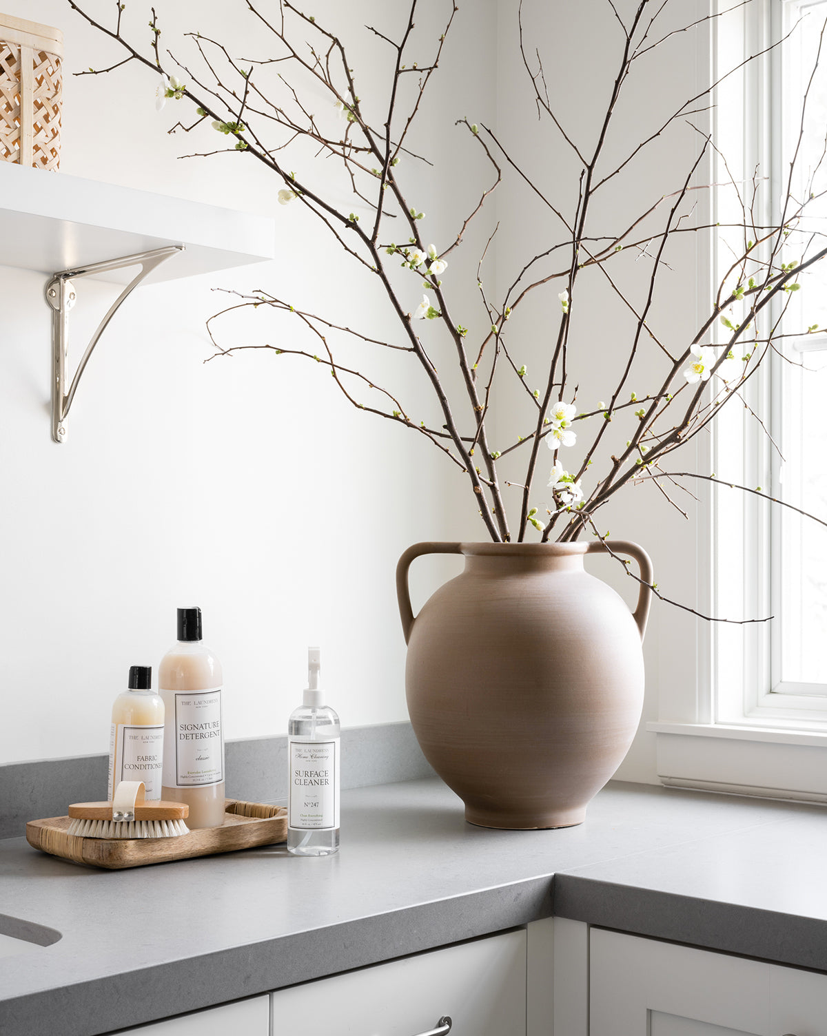 A Rounded Dual Handled Vase with long branches and white blossoms sits on a gray kitchen countertop near a window, next to soap and cleanser bottles neatly arranged on a wooden tray.