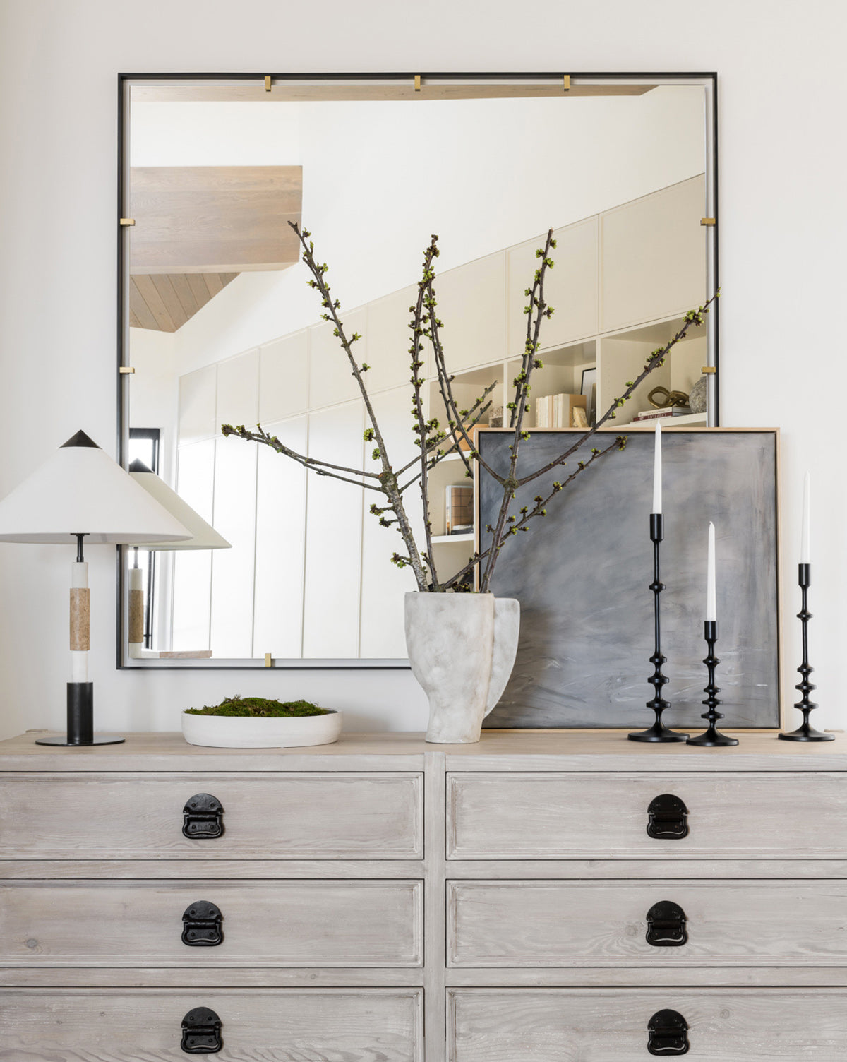 A light wood dresser displays the Quiet artist-enhanced print, a modern table lamp, a moss bowl, a white branch vase, and black candleholders. Above, a square mirror reflects tonal grays in the bright, minimal room.