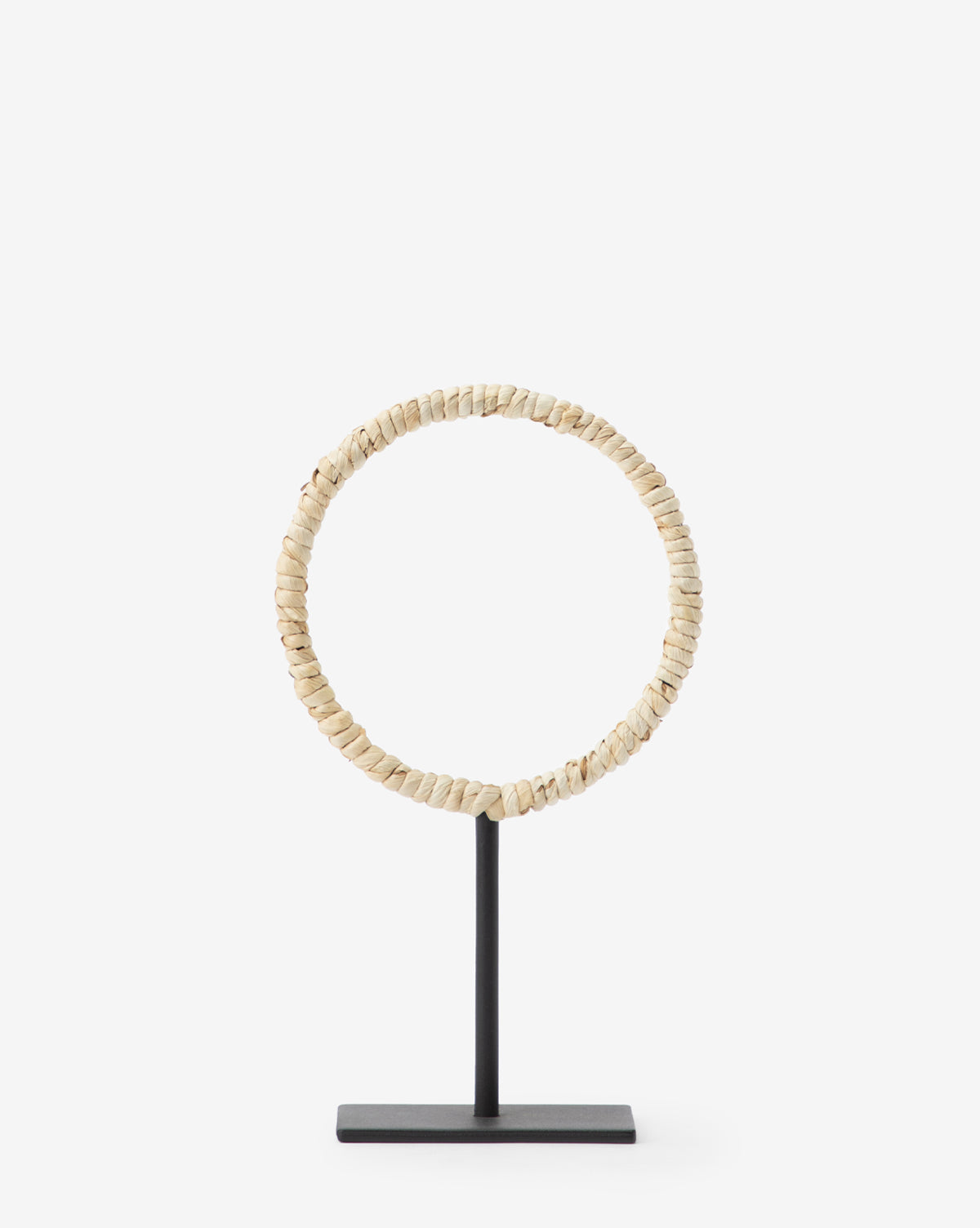The Brayden Woven Object is a circular necklace of small, cream-hued beads with a corn husk texture, displayed upright on a slim black metal stand with a rectangular base—ideal for contemporary decor against a plain white background.