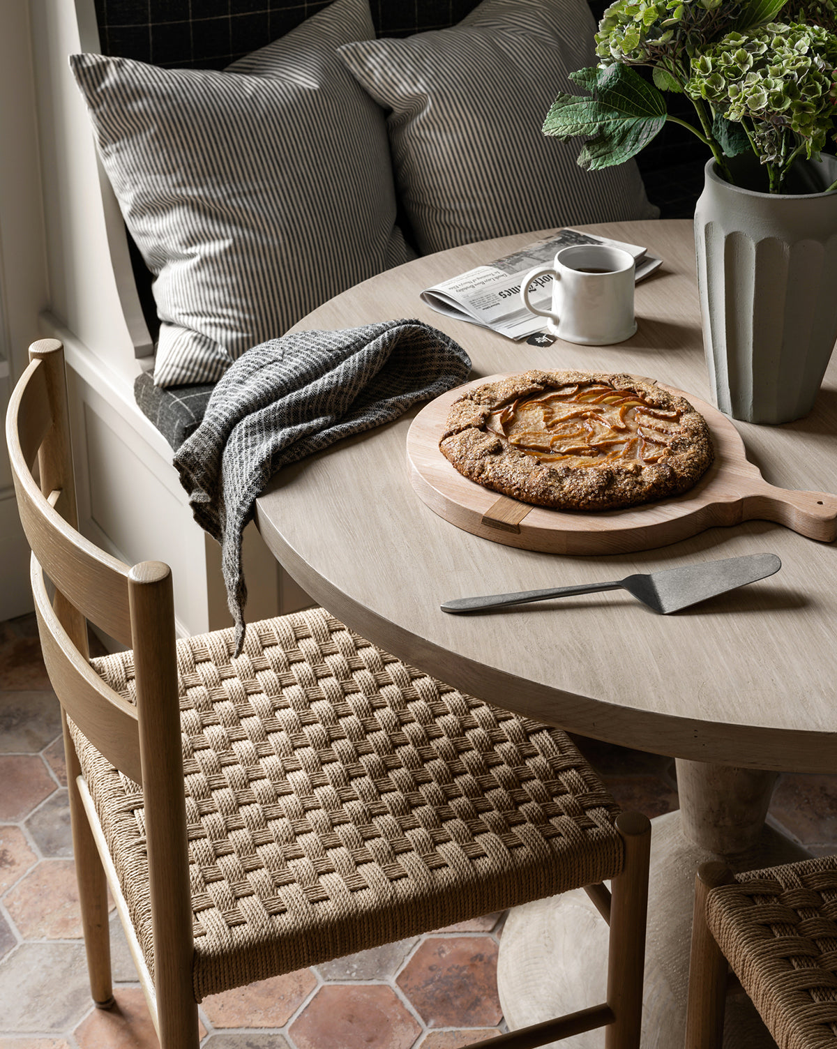 A cozy breakfast nook features Danish design with Eloise Woven Chairs and a round wooden table topped with a rustic fruit galette, pie server, mug, newspaper, and green hydrangeas. Pillows and a blanket add comfort, by McGee & Co.