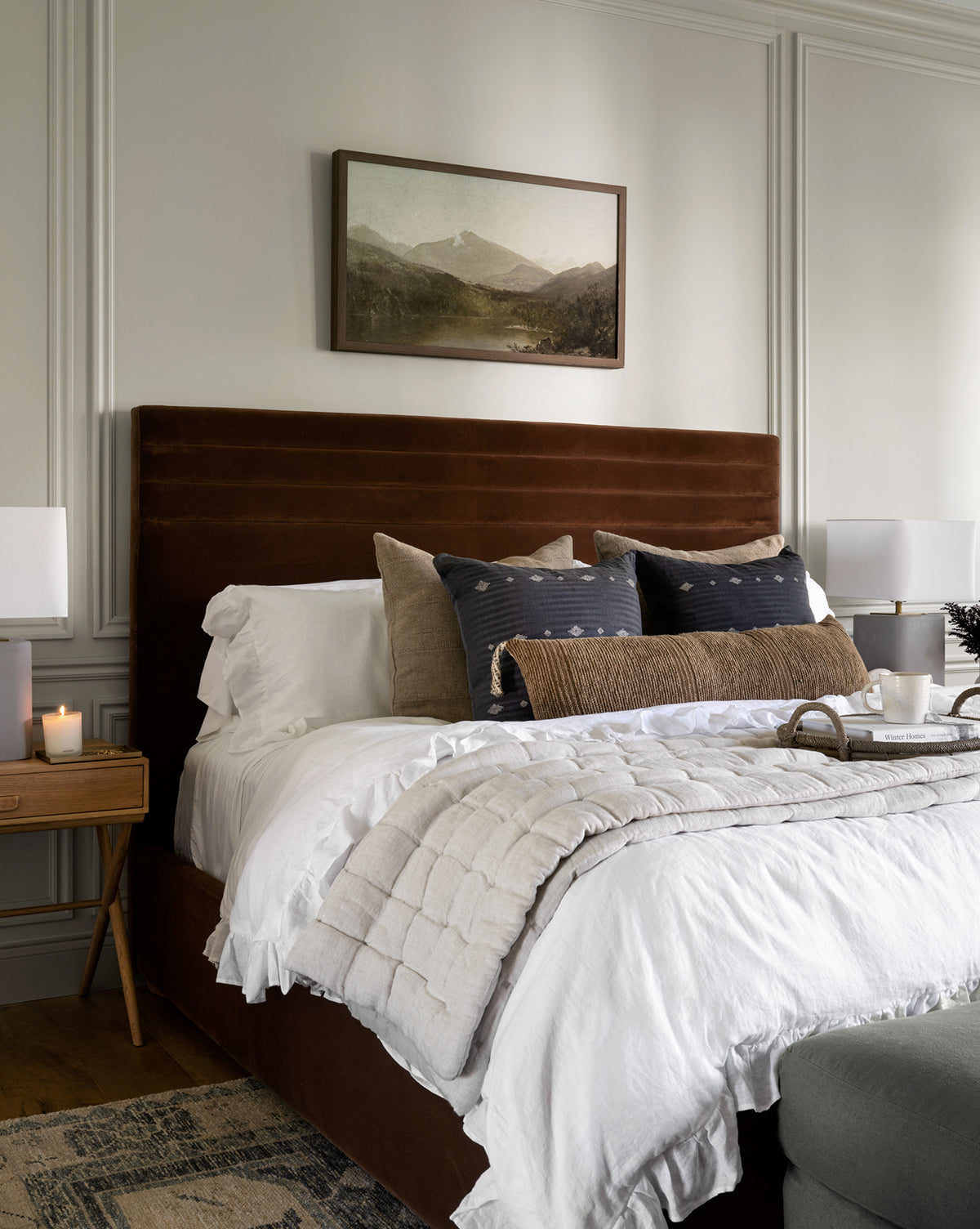 A cozy bedroom features a brown upholstered bed with white bedding, neutral pillows, and the Chaucer Quilt in gray adding textured style. Nightstands display lamps and candles, while a tray with cups is set on the bed.