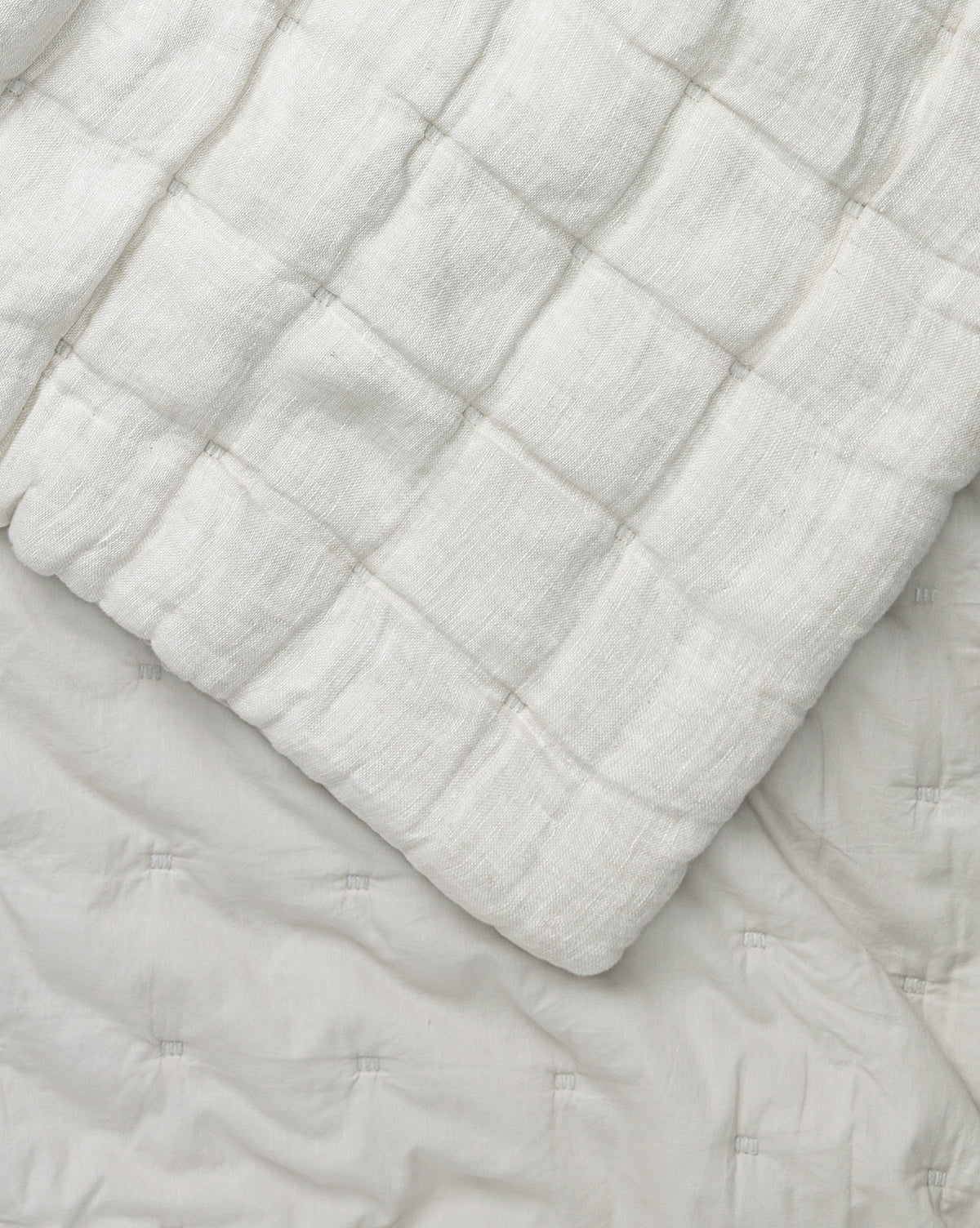 A close-up of the Chaucer Quilt reveals its soft, white fabric and grid-patterned quilted stitching, draped over a matching white linen and cotton surface with tufted details.
