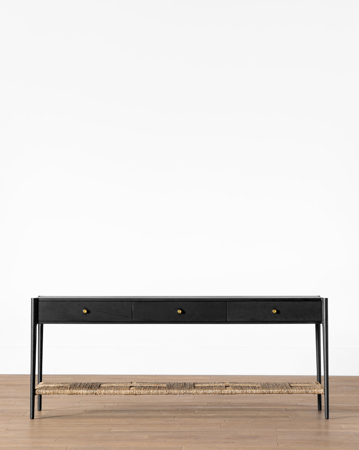 The Dana Console is a minimalist black table featuring three drawers with gold knobs, slim legs, and a woven seagrass shelf—perfect entryway decor against a white wall on a wooden floor, by McGee & Co.