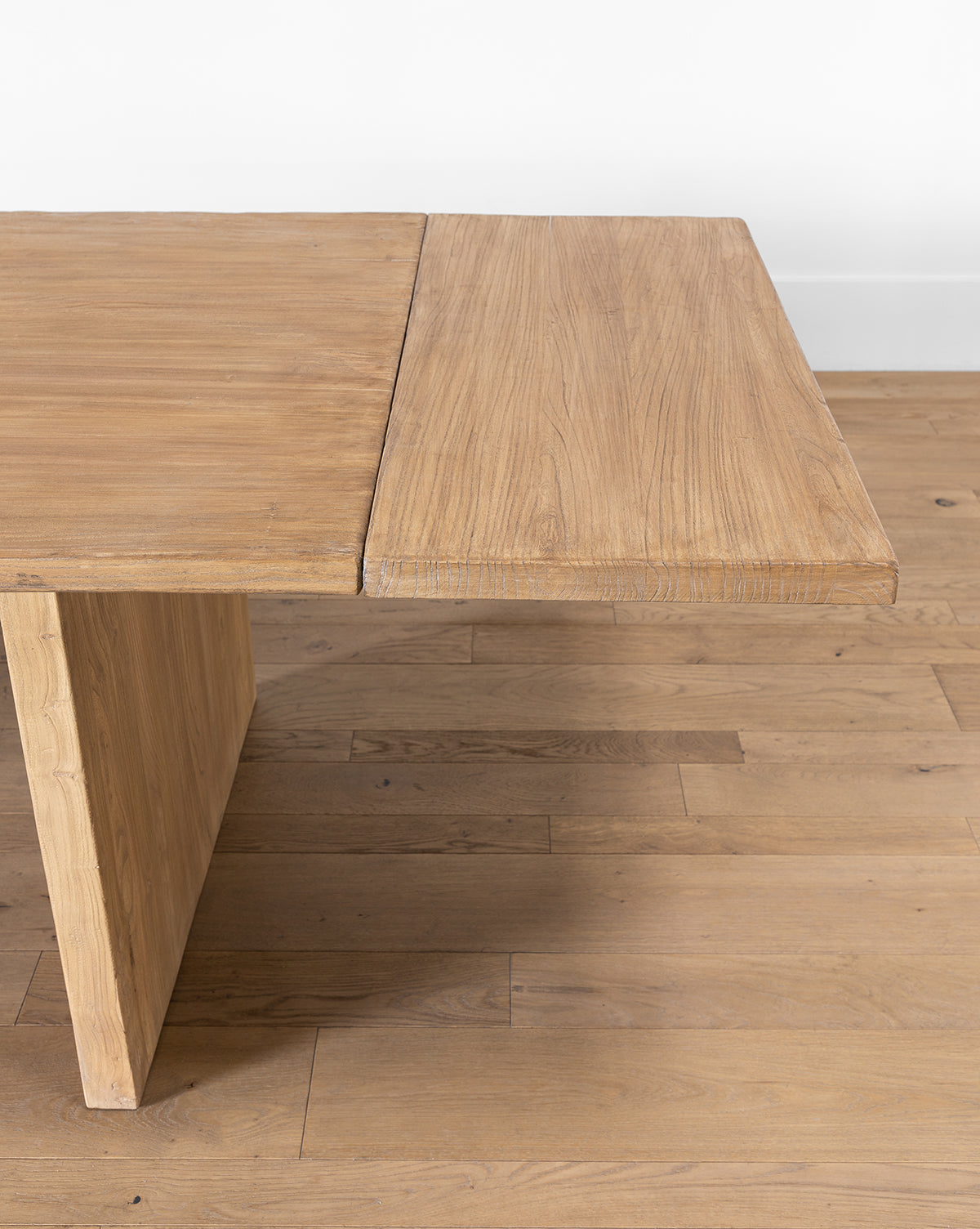 A close-up of the Dillon Extension Dining Table shows its rectangular shape, thick textured top, and angled leg, set on a wooden floor against a white wall by McGee & Co.