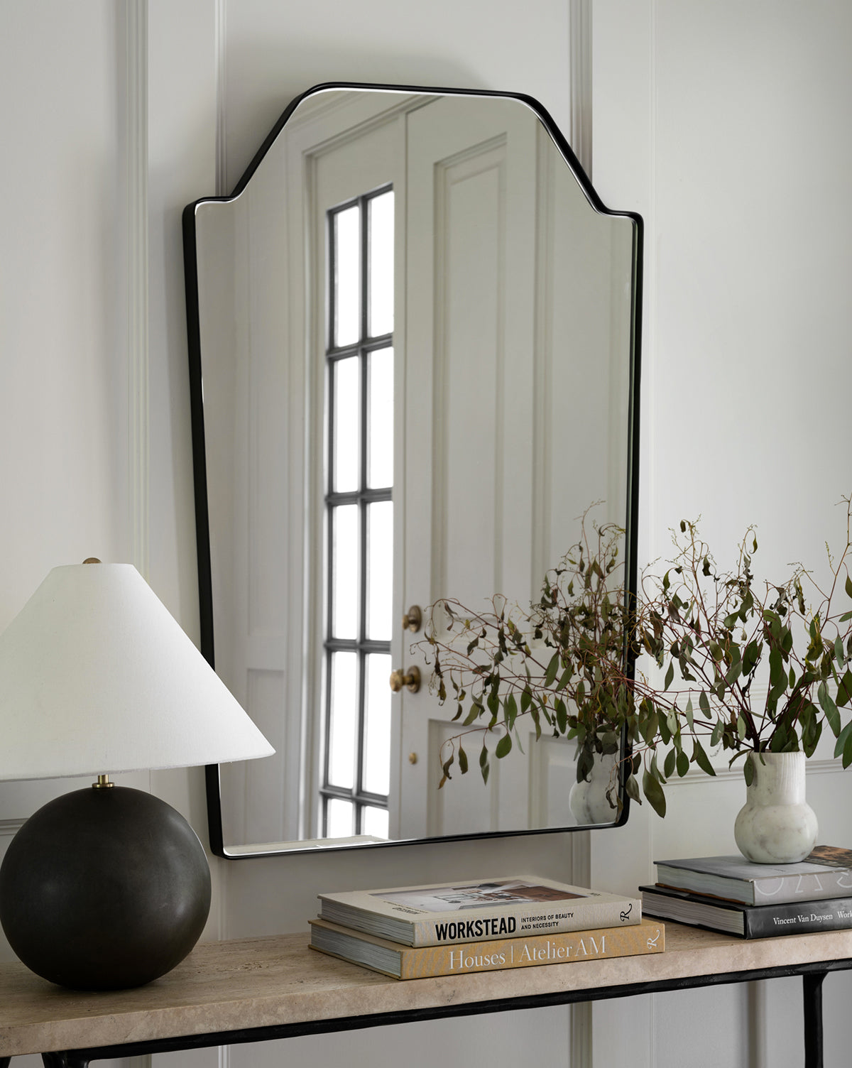The Giles Wall Mirror with its unique shape hangs above a console table holding a matte black lamp, stacked books, and a vase with leafy branches, while the mirror reflects a door with glass panels by McGee & Co.