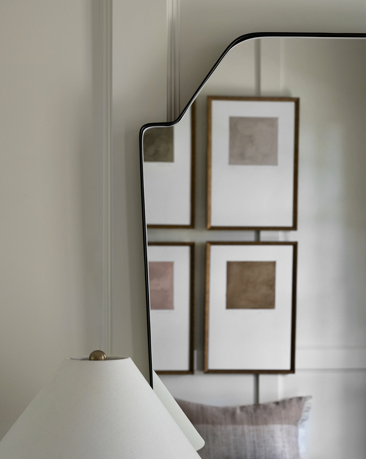 The Giles Wall Mirror reflects four framed abstract artworks, a white lamp with a gold finial, and part of a gray pillow, all set against neutral-toned walls and décor, by McGee & Co.