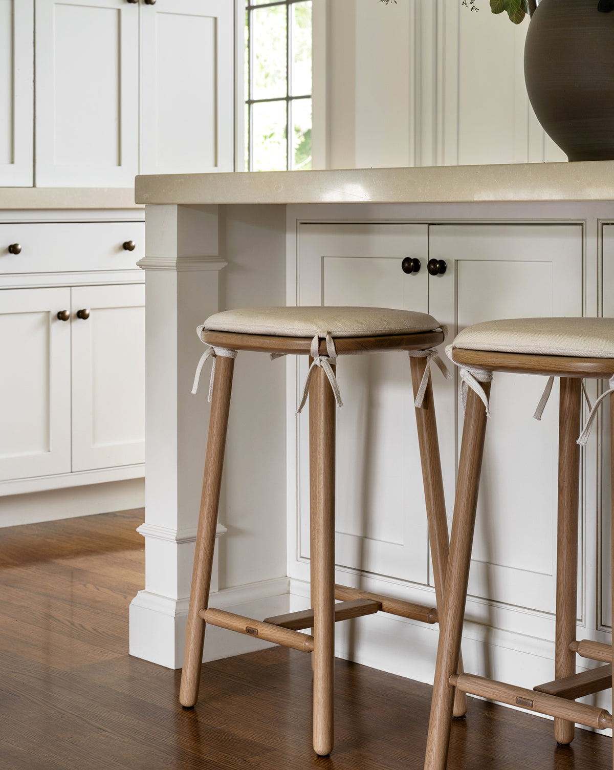 Two Ralph Oak Counter Stools with linen cushions sit at a kitchen counter featuring white cabinetry and a beige countertop, while sunlight through a window highlights the warm wood flooring, by McGee & Co.
