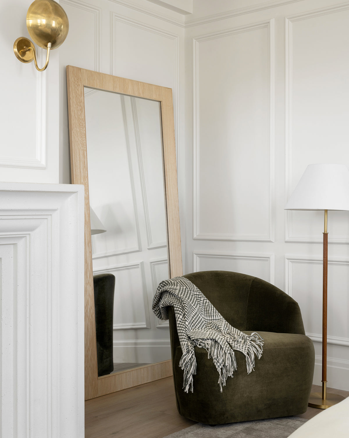 A modern corner with a green velvet Byron Chair draped with a patterned throw, a large wooden-framed mirror, a white floor lamp, white paneled walls, and a brass wall sconce, by McGee & Co.