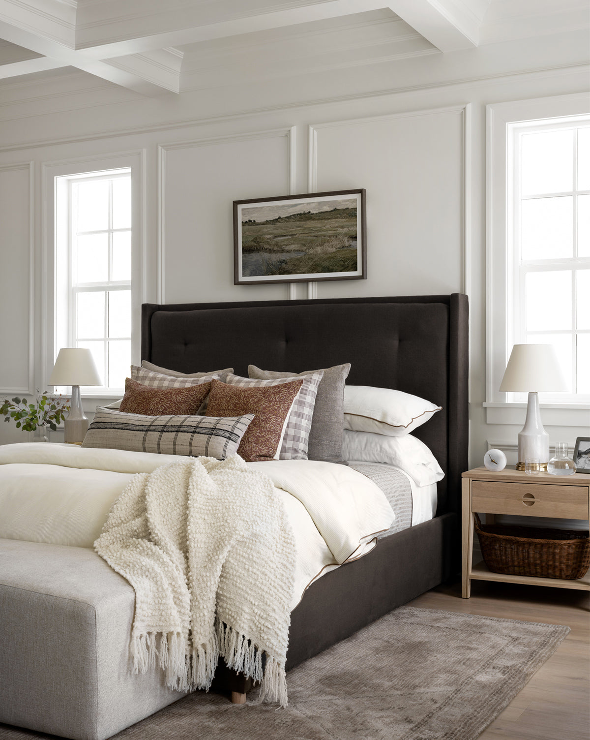 The Ria Bed by Makerspalm, topped with layered pillows and a cream knit blanket, sits in a bright modern bedroom with white paneled walls, large windows, dual bedside tables, and a landscape painting above its tufted headboard by McGee & Co.