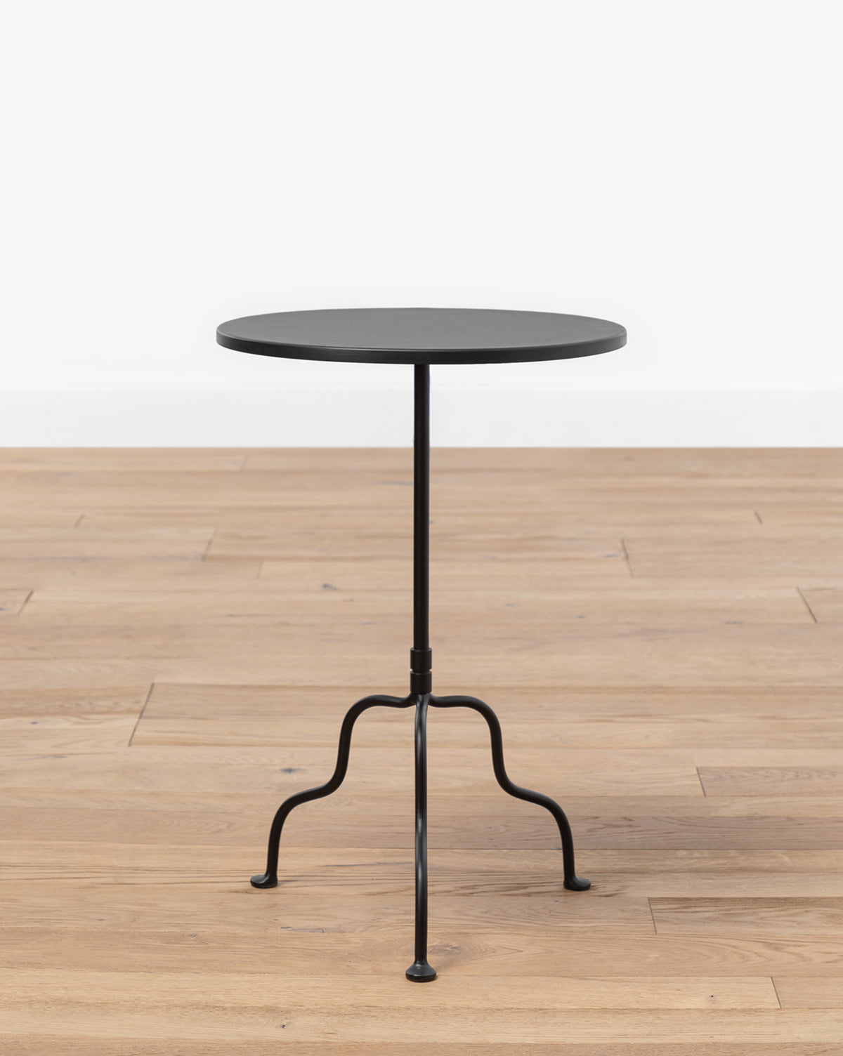 The Goodrich Iron Side Table, featuring a small round matte black top and slender three-legged base, stands on a light wood floor against a plain white background by McGee & Co.