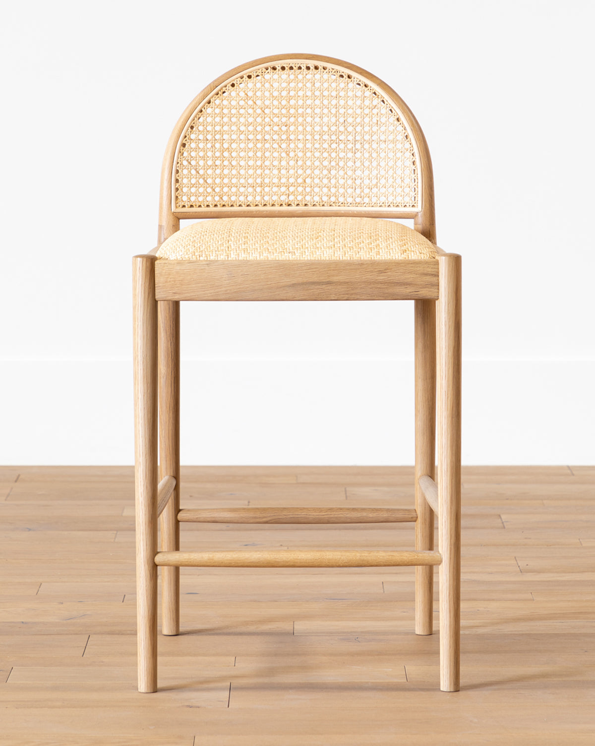The Hadden Natural Counter Stool features an oak frame, woven rattan seat and backrest, rounded back, and straight legs—ideal for coastal décor—shown here on a light wooden floor against a plain white wall by McGee & Co.