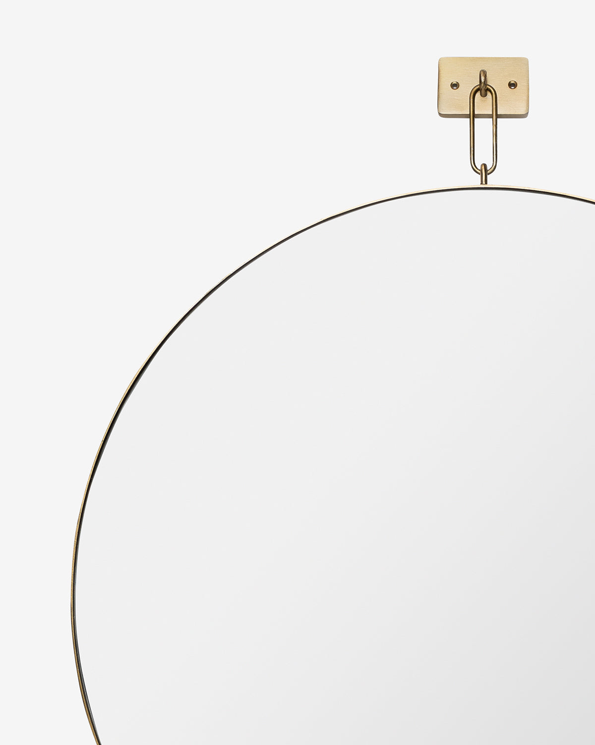The Haven Wall Mirror, featuring a thin stainless steel frame, hangs from a gold wall mount and chain against a plain white background. Only the top part of this modern, elegant mirror is shown in the image, by McGee & Co.