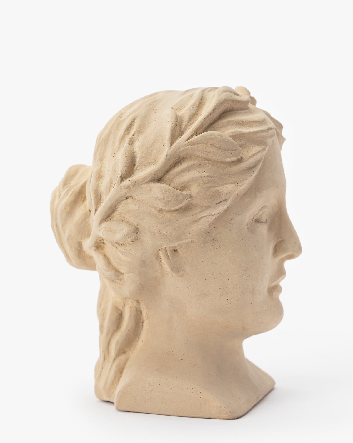 The Hestia Bust: a beige composite stone sculpture of a woman with wavy hair, tied back and adorned with a laurel wreath, shown in profile facing right. An elegant artistic decor piece for a sophisticated setting.