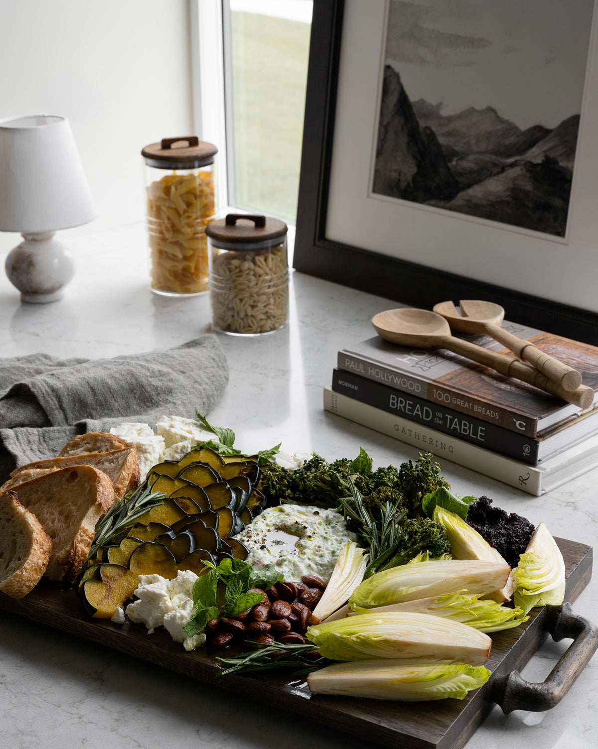 A charcuterie board with grilled squash, broccoli, nuts, cheese, and endive is served on the Fordham Board near toasted bread, books, wooden utensils, pasta jars, a lamp, and framed art by the window by McGee & Co.